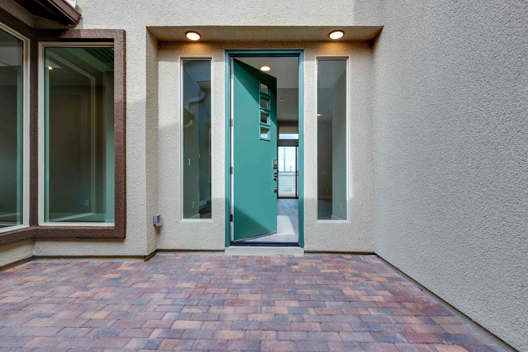 The image shows a modern entryway with a teal-colored glass door, surrounded by windows and a paved brick walkway leading up to the entrance.