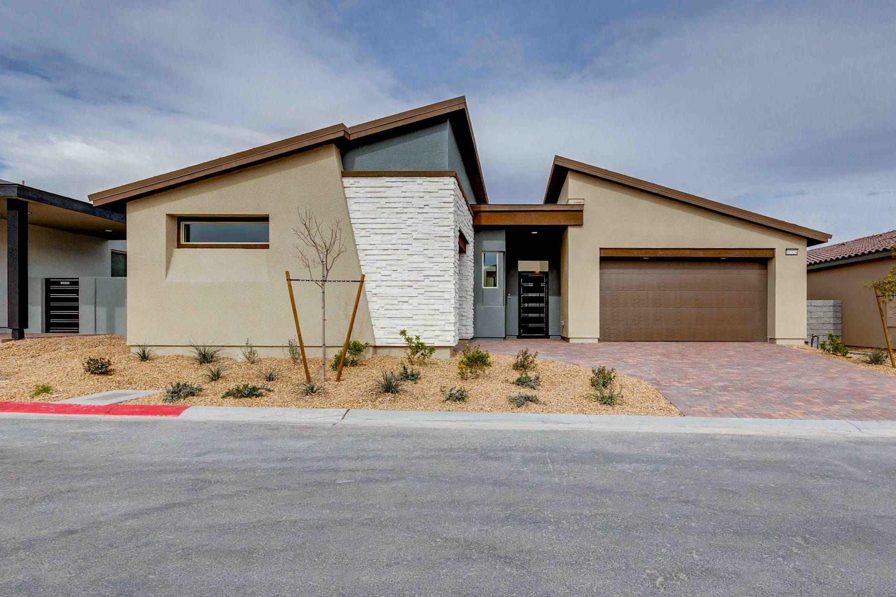 A modern, single-story house with a mix of beige and white exterior, a garage door, and a paved driveway in the foreground, set against a blue sky with scattered clouds.