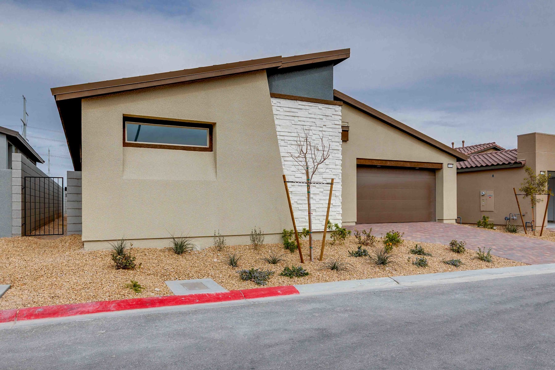 A modern, single-story house with a garage, surrounded by a gravel driveway and landscaped with small plants and trees against a cloudy sky.