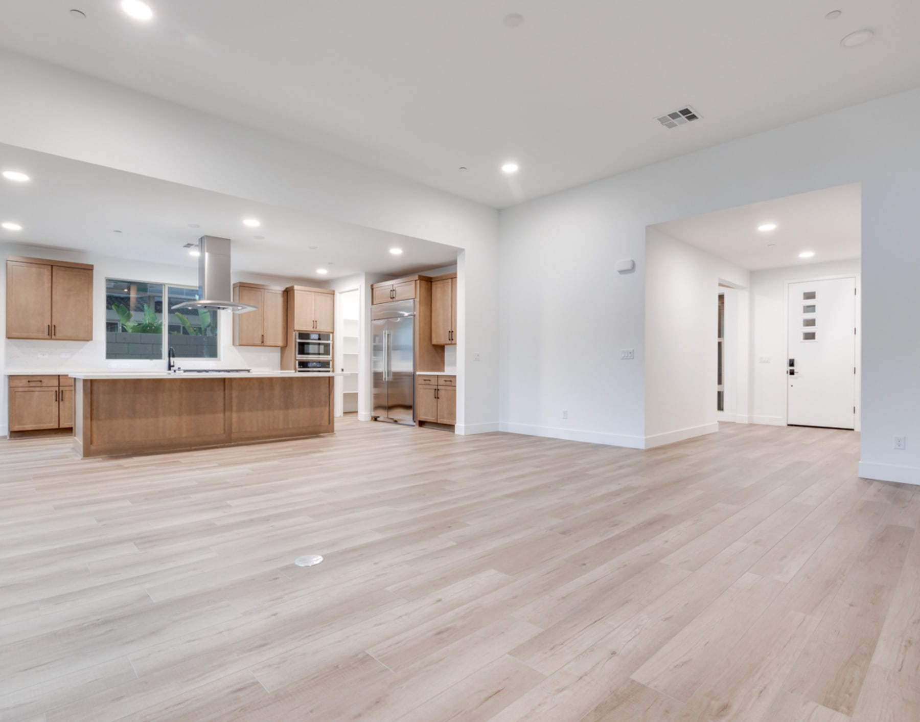 A spacious, open-concept kitchen and living area with light-colored hardwood floors, white walls, and modern wooden cabinetry.