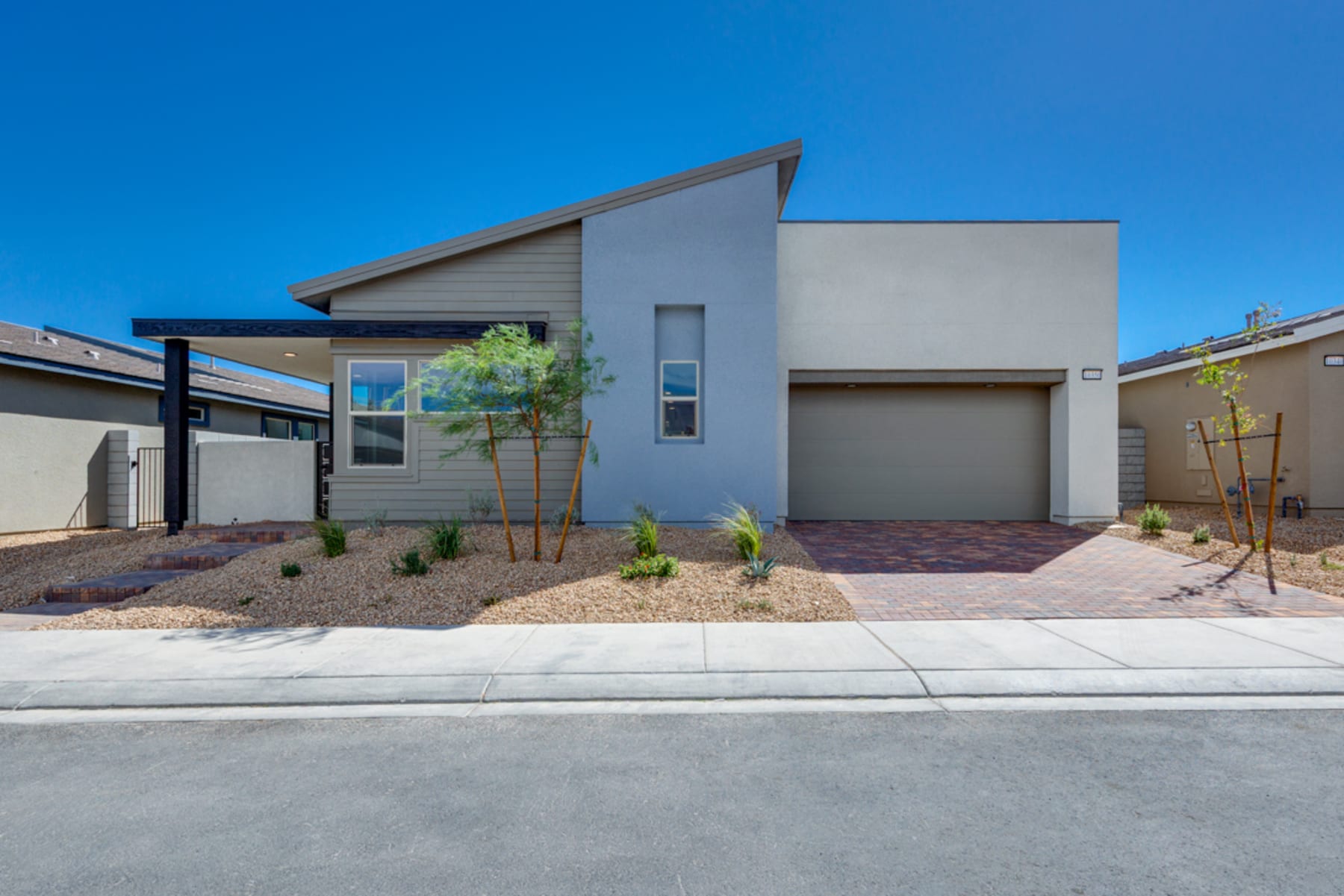 A modern, single-story residential home with a gray exterior, a slanted roof, and a well-landscaped front yard featuring desert-adapted plants against a clear blue sky.