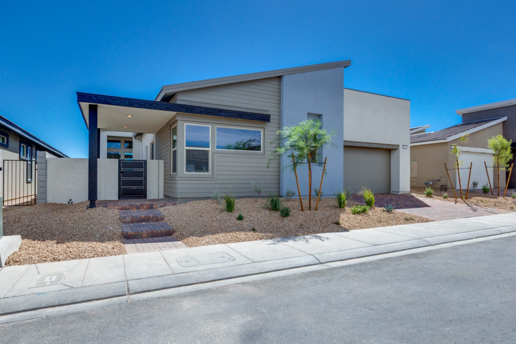 A modern, single-story residential home with a gray exterior, a blue roof, and a well-landscaped front yard featuring desert-adapted plants and a paved walkway leading to the entrance.