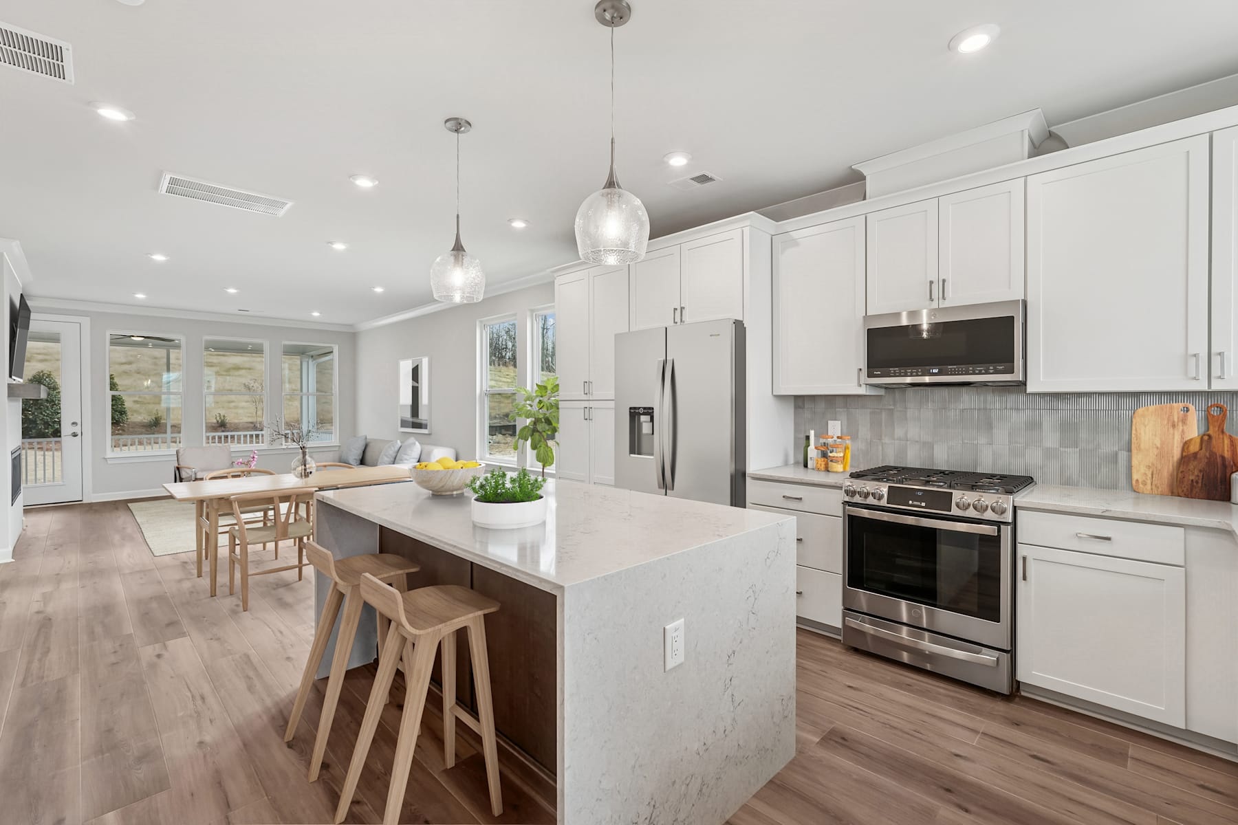 A modern, open-concept kitchen with white cabinets, stainless steel appliances, and a large island with bar stools, set against a backdrop of hardwood floors and pendant lighting.