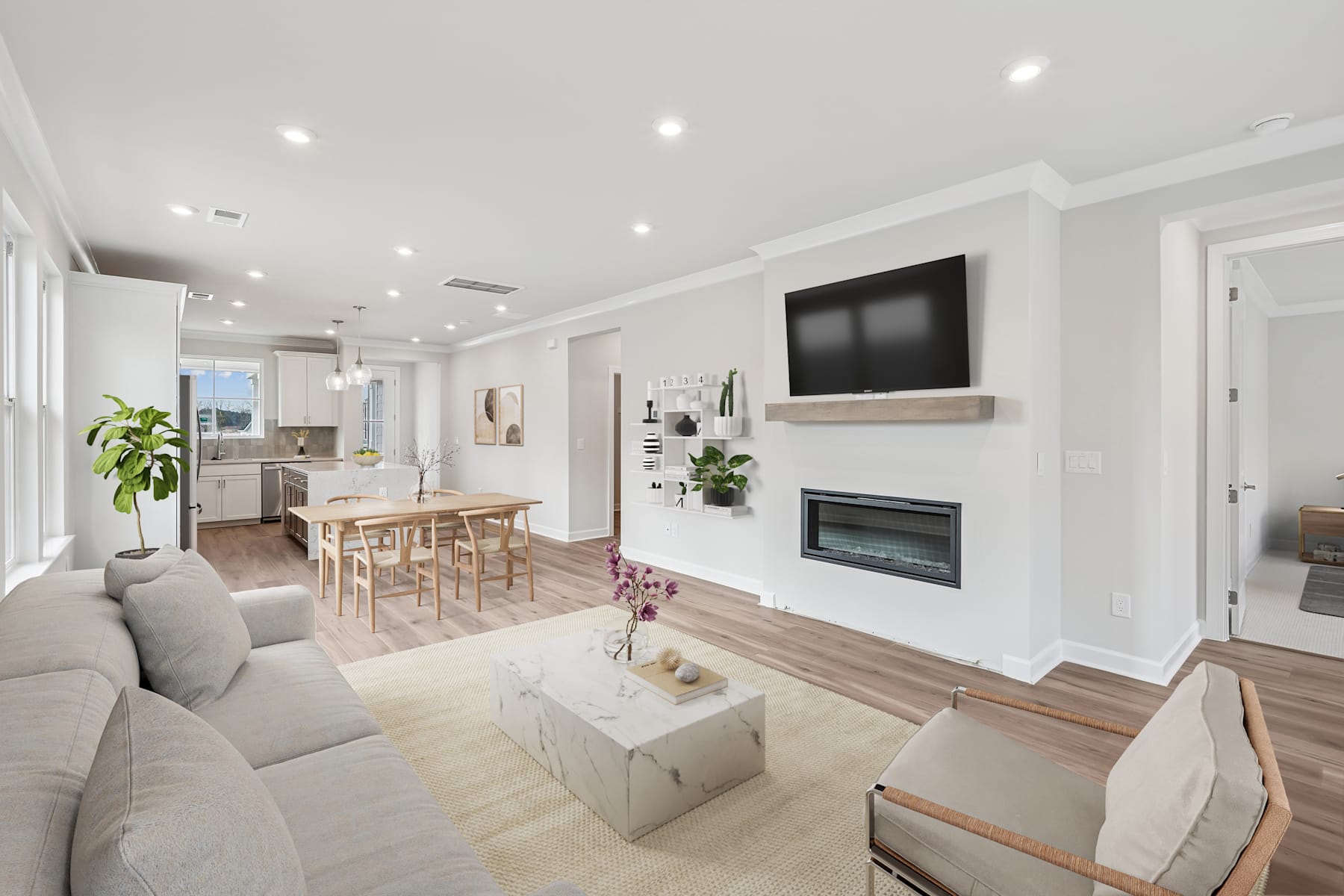 A modern, open-concept living room with a fireplace, a large TV mounted on the wall, and a dining area visible in the background, all furnished with neutral-toned furniture and decor.