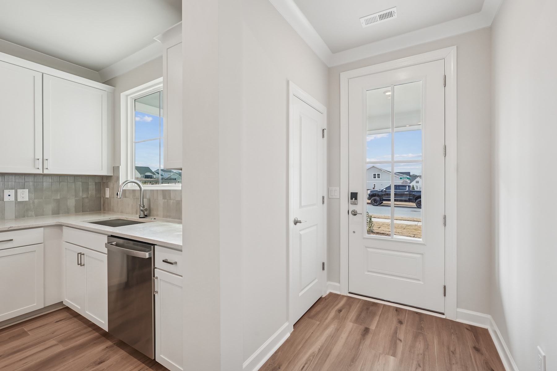A modern, bright kitchen with white cabinets and appliances, leading to a hallway with a glass door providing a view of the exterior.