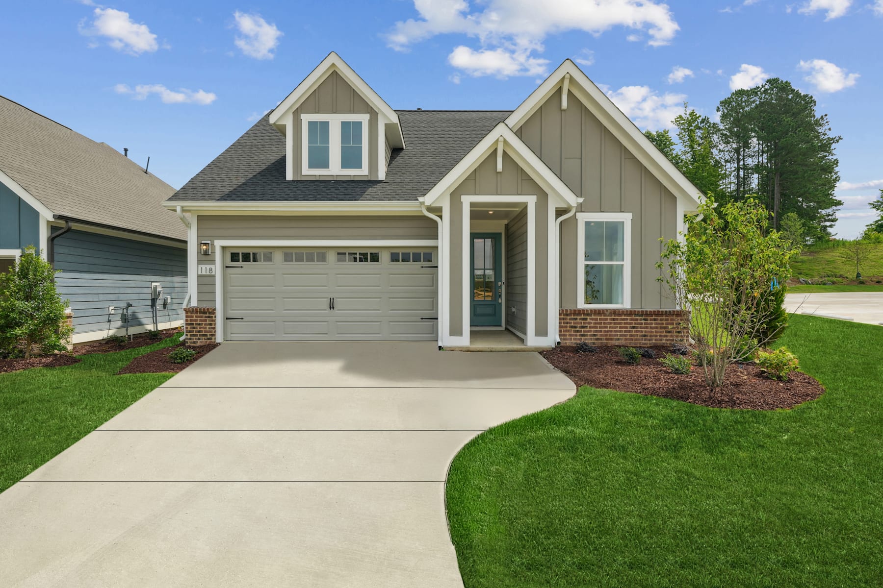 A well-manicured lawn leads to a two-story house with a gray exterior, a garage, and a lush green landscape in the background.