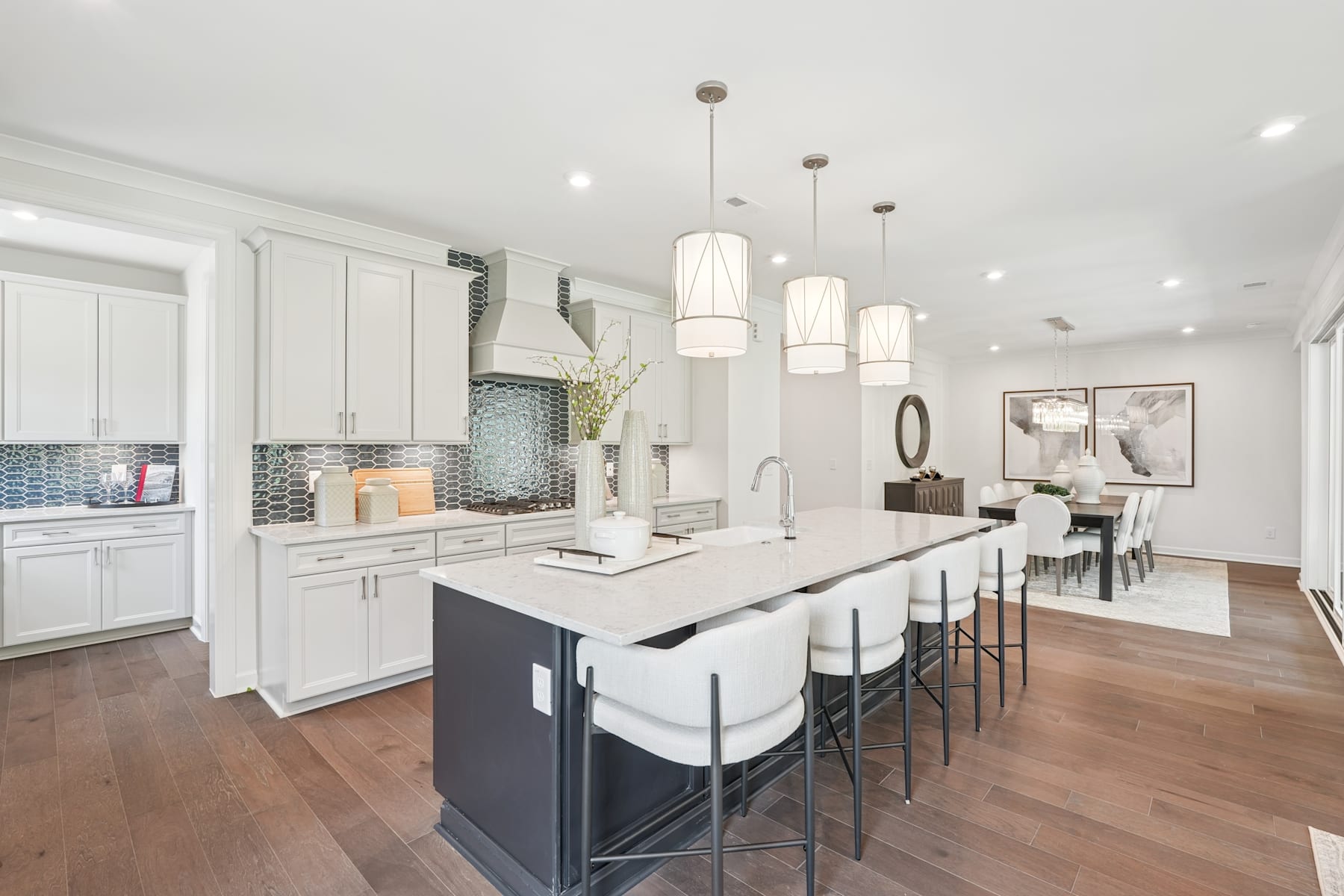 A modern, open-concept kitchen with white cabinets, a large island with bar stools, and pendant lighting fixtures, leading into a dining area with a wooden table and chairs.