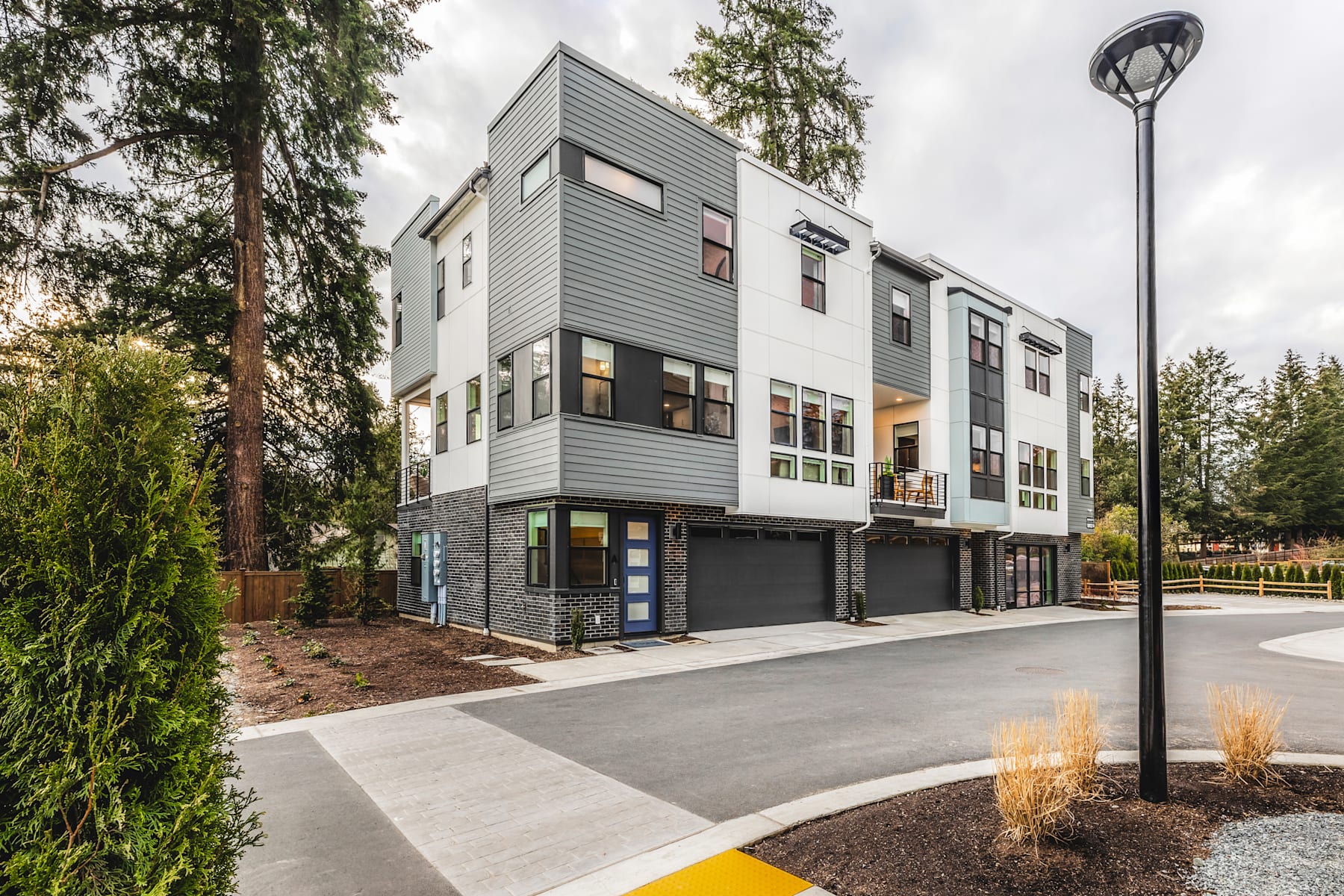 A modern, multi-story residential building with a gray exterior stands amidst a landscaped environment featuring trees, shrubs, and a paved walkway in the foreground.