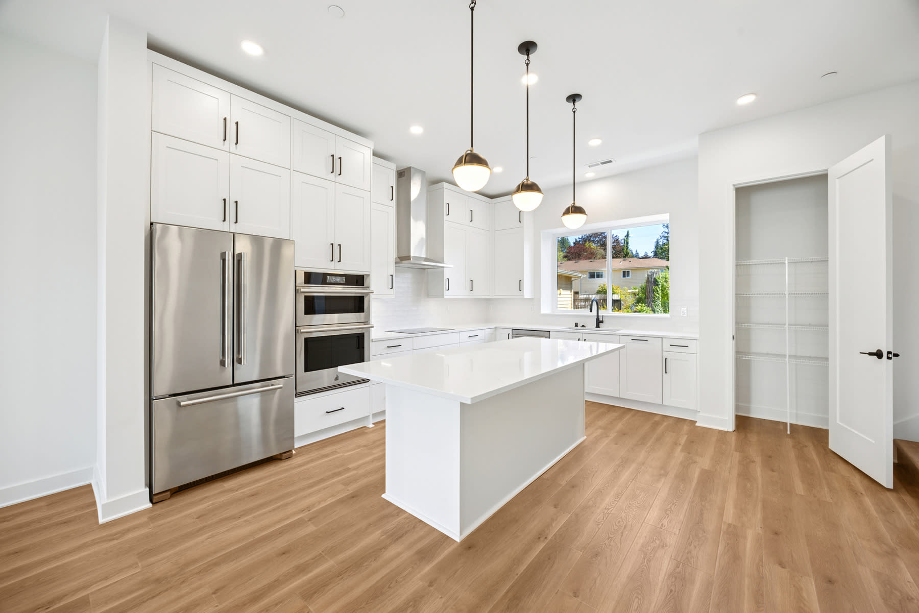 A modern, bright kitchen with white cabinets, stainless steel appliances, and a wooden floor, with a window providing natural light and a view of the outdoors.
