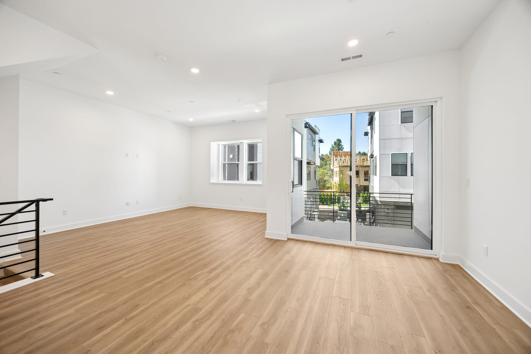 A bright, spacious room with hardwood floors, large windows, and a balcony overlooking a residential neighborhood.