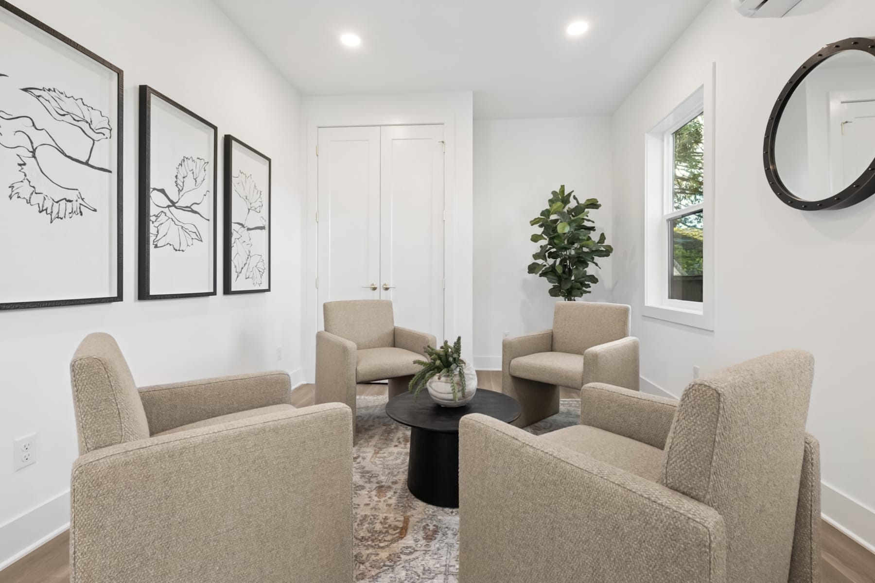 A cozy and modern living room with beige armchairs, a round coffee table, and framed artwork on the walls, complemented by a large window providing natural light.