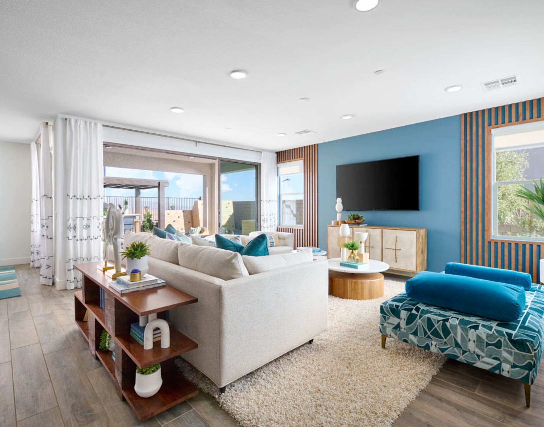 A cozy and modern living room with a white sofa, a blue patterned armchair, and a wooden entertainment unit with a large TV mounted on the wall. The room features large windows that let in natural light and provide a view of the outdoor area.