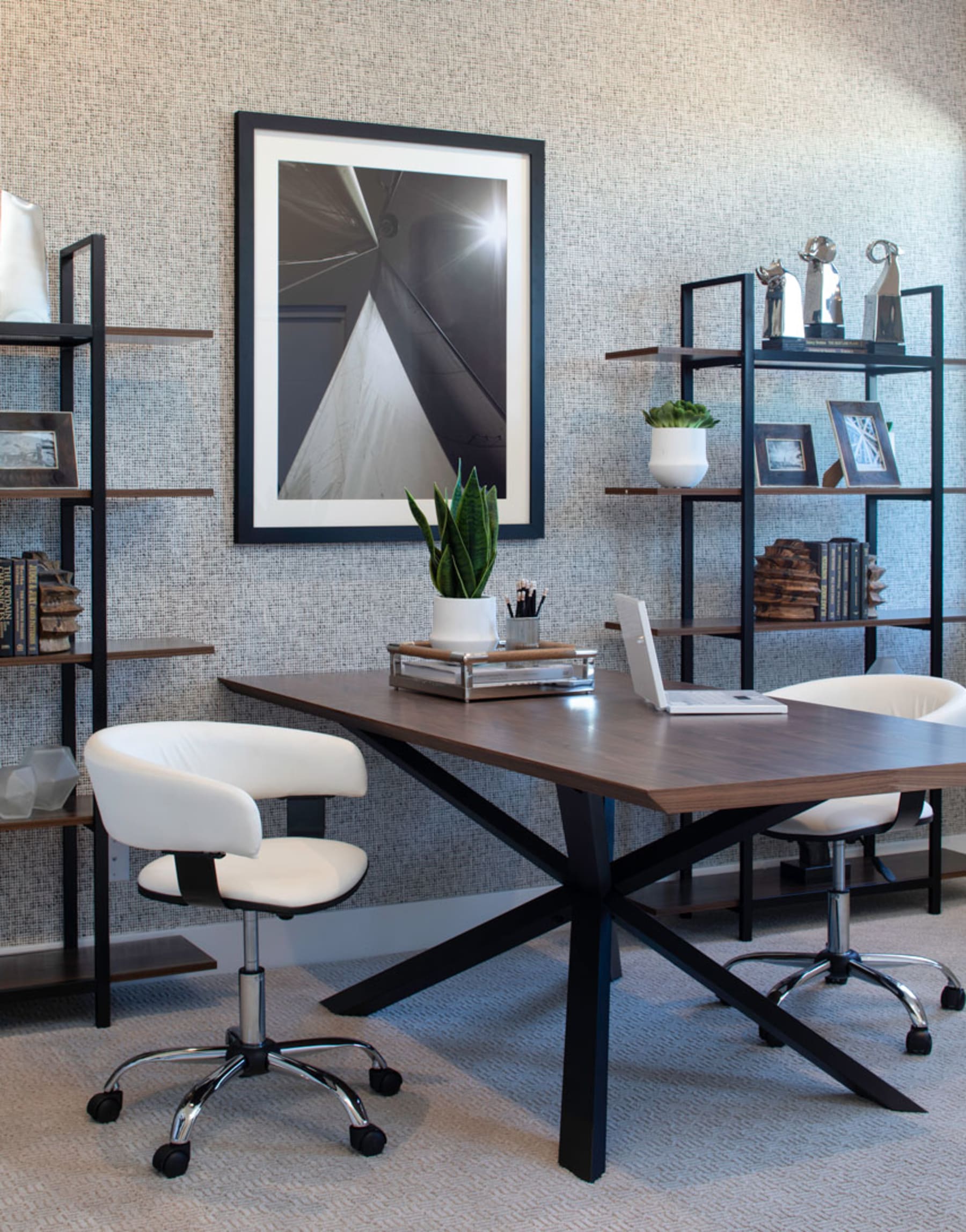 A modern and minimalist home office space with a wooden desk, black metal frame, and various decorative elements such as shelves, plants, and framed artwork on the wall.