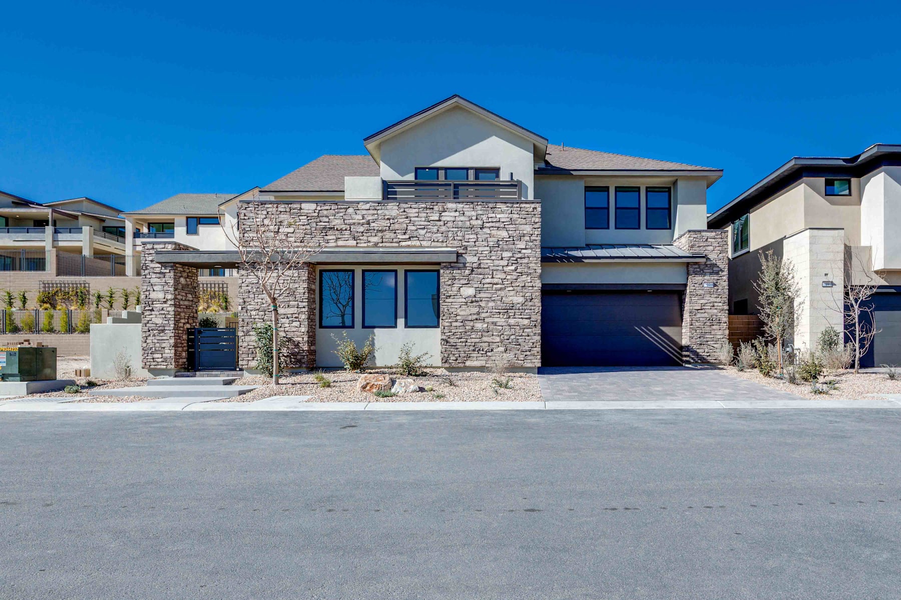 A modern, two-story house with a stone exterior, large windows, and a garage door stands in the foreground, set against a clear blue sky in the background.
