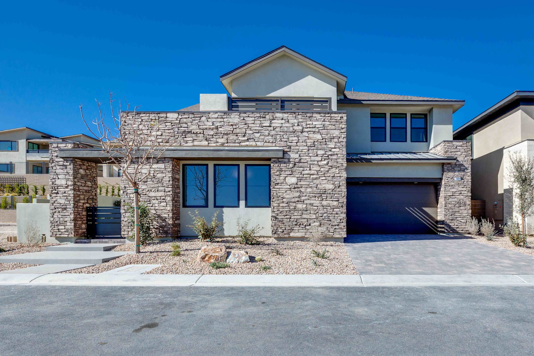 A modern, two-story house with a stone exterior, large windows, and a garage door stands in the foreground, set against a clear blue sky in the background.