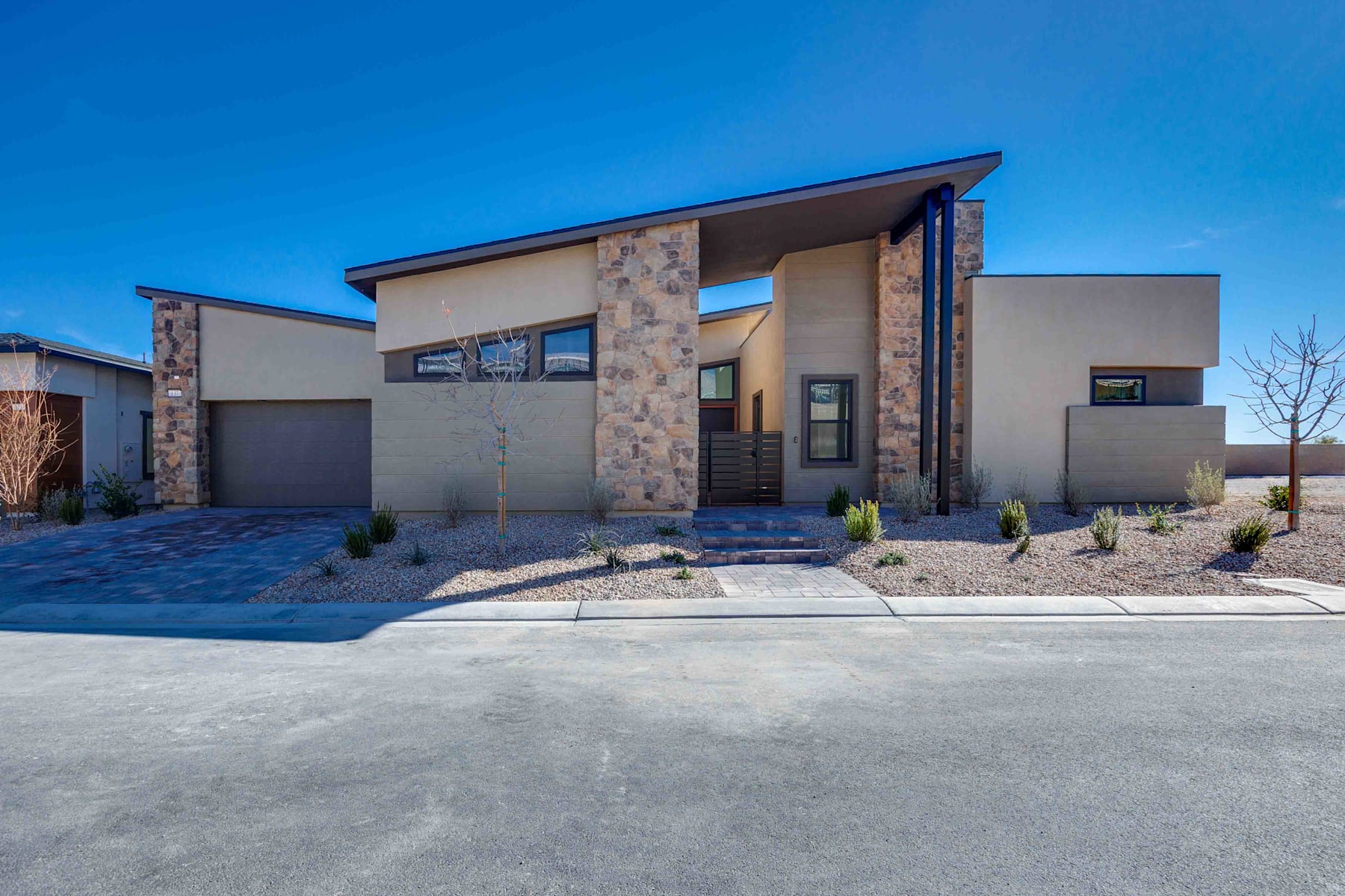 A modern, two-story residential home with a mix of stone and stucco exterior, surrounded by a paved driveway and landscaped yard against a clear blue sky.