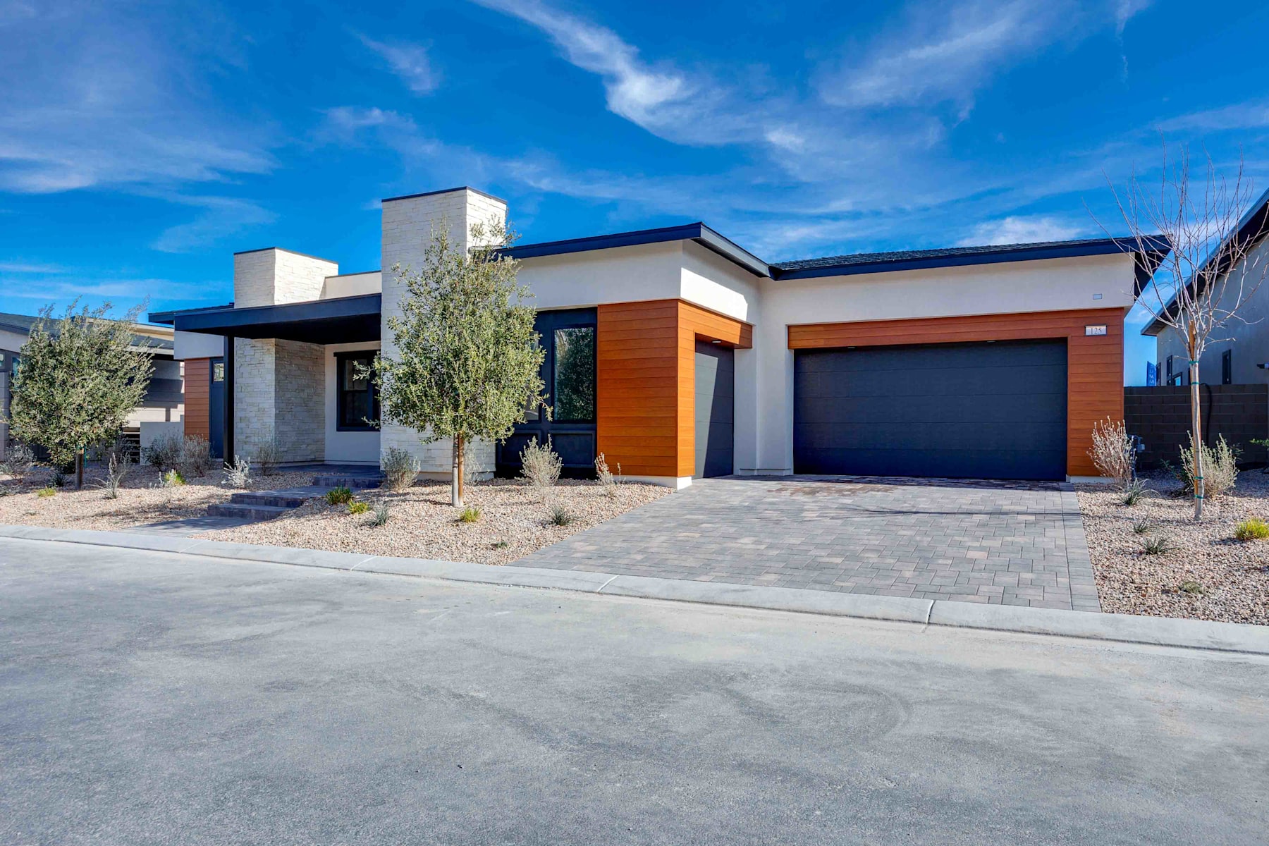 A modern, single-story house with a garage, surrounded by a paved driveway and landscaped with desert vegetation against a backdrop of a clear blue sky with wispy clouds.
