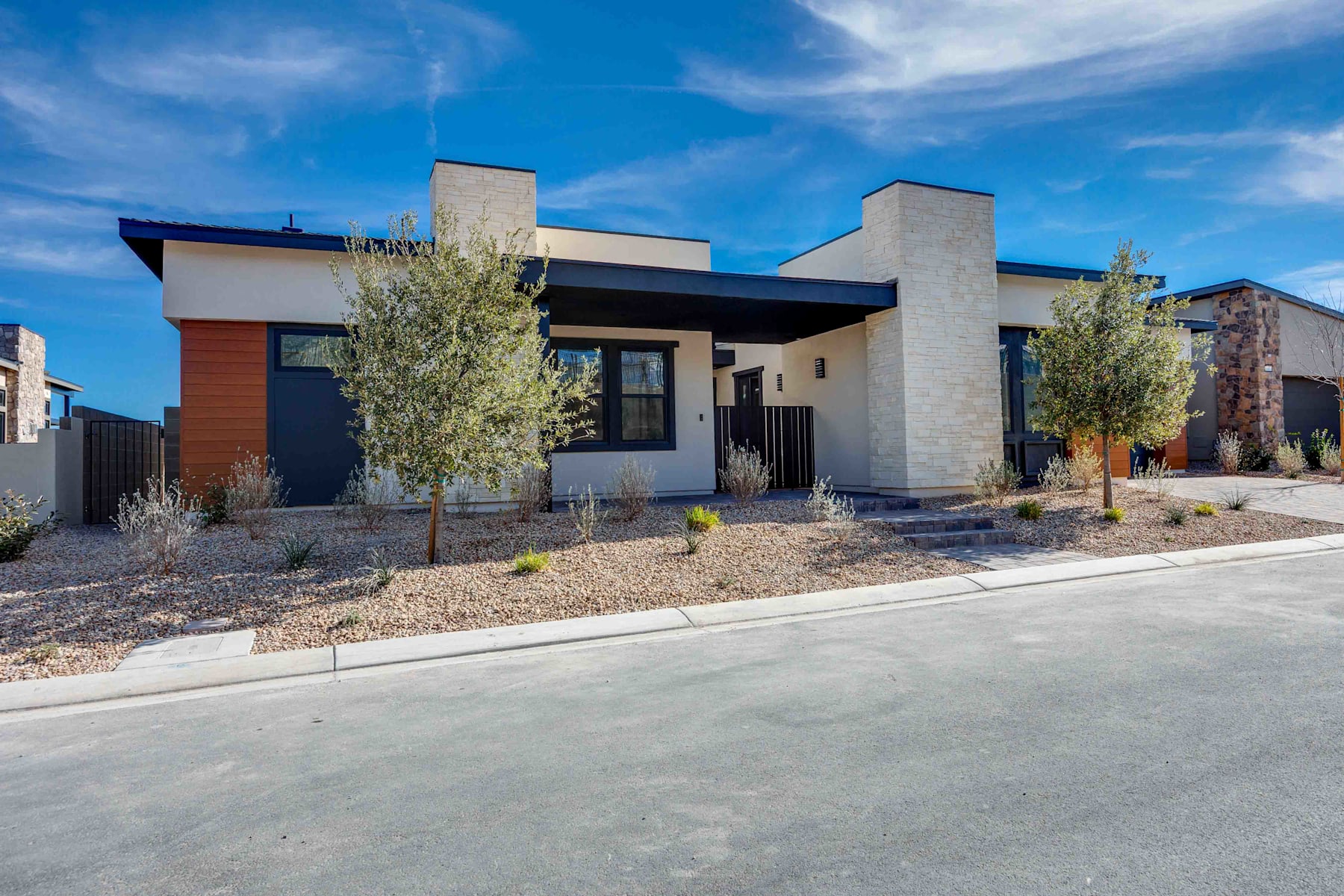 A modern, single-story residential building with a mix of materials, including stone and wood, surrounded by landscaping and situated against a clear blue sky.