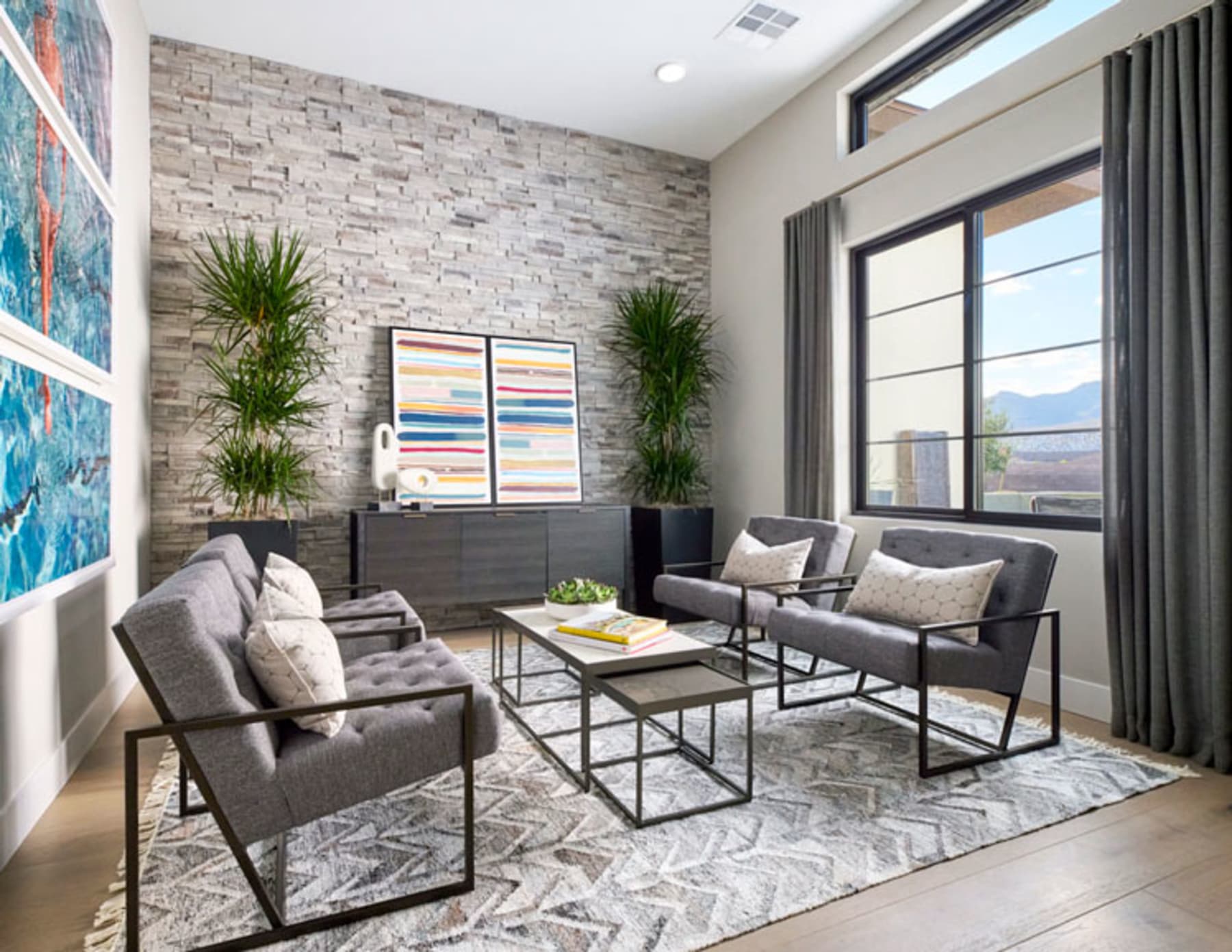 A modern and stylish living room with a gray sectional sofa, a glass coffee table, and a stone accent wall with potted plants, complemented by large windows offering a scenic view.