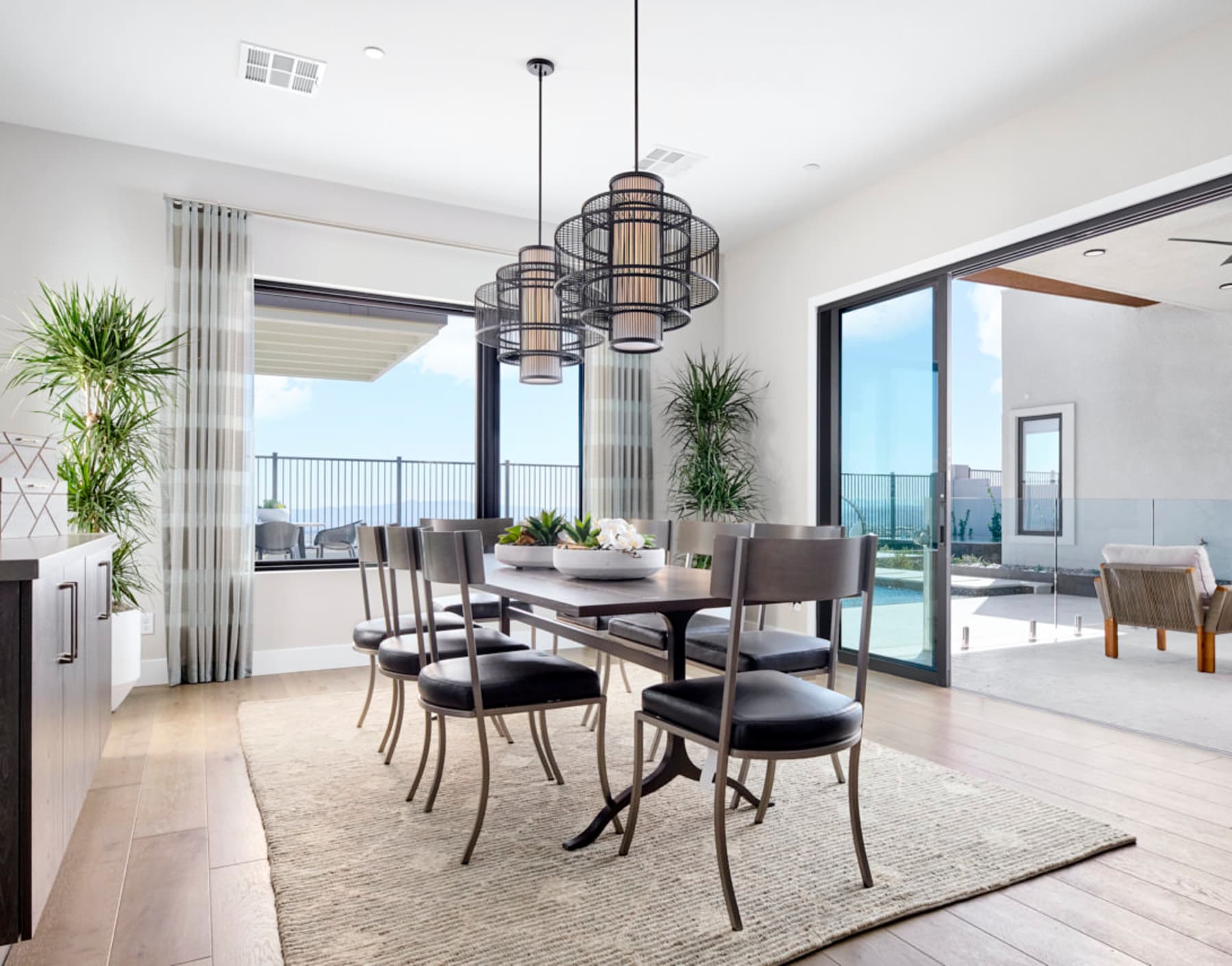 A modern and spacious dining room with a large wooden table, comfortable chairs, and stylish pendant lights, overlooking a scenic outdoor view through the large windows.