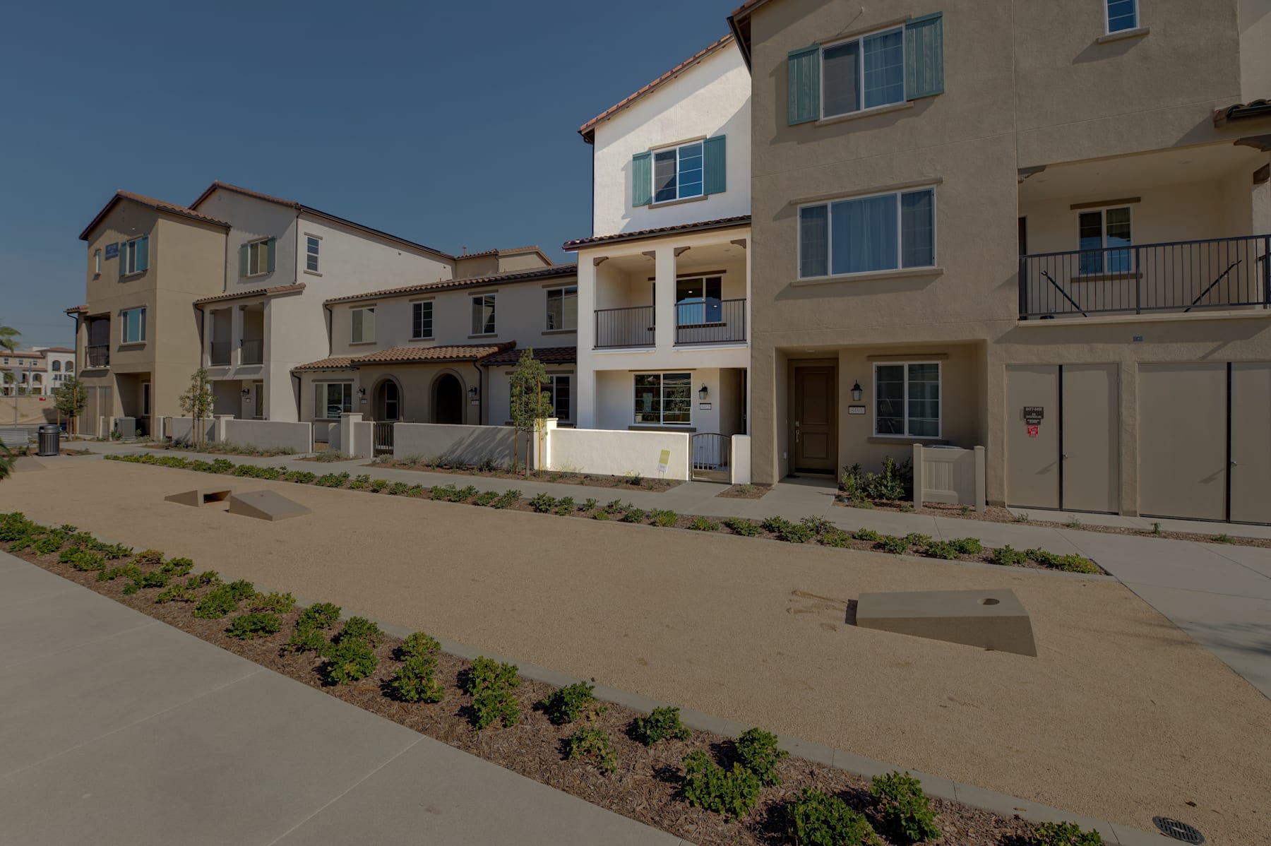 A row of multi-story residential buildings with a paved walkway and landscaped greenery in the foreground, set against a clear blue sky.