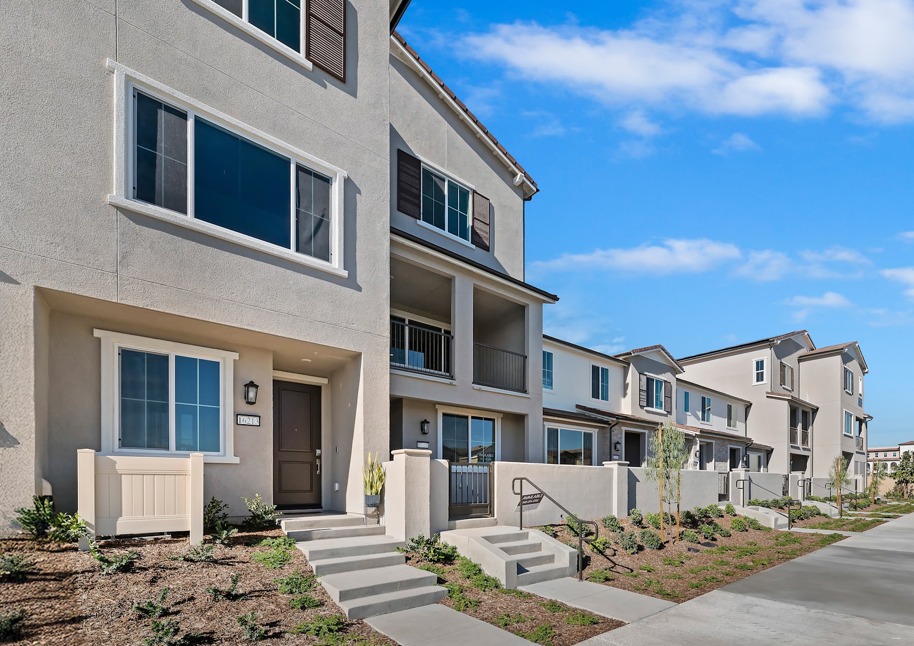 A row of modern, multi-story townhouses with gray exteriors and blue-tinted windows, set against a backdrop of a bright, cloudy sky.