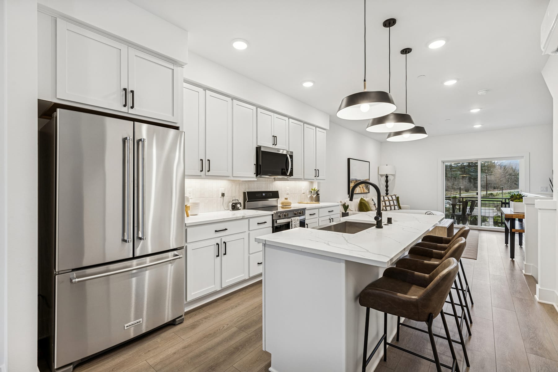 A modern, well-lit kitchen with white cabinets, stainless steel appliances, and a central island with bar stools, set against a backdrop of hardwood floors and large windows.
