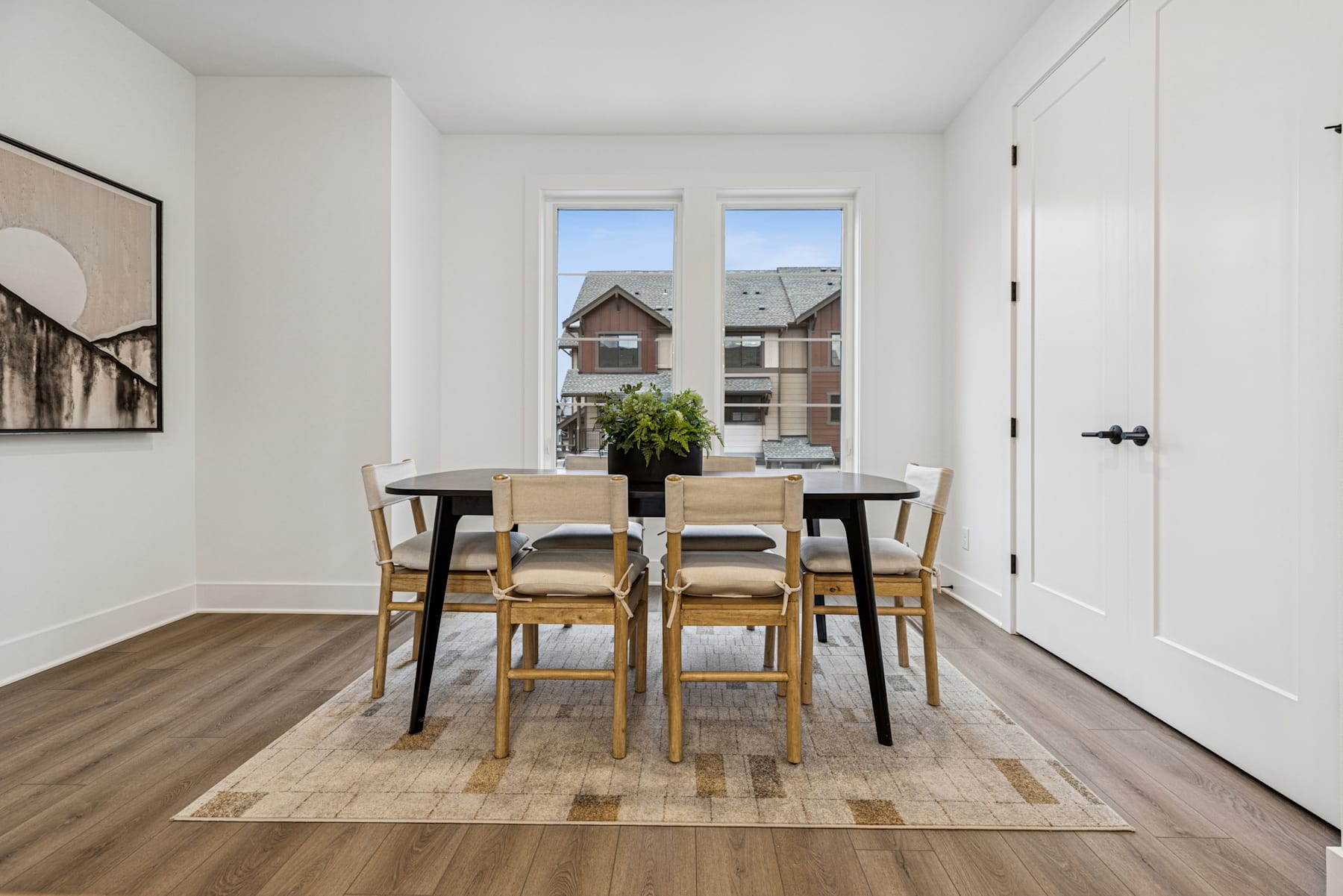 A modern and minimalist dining area with a wooden table and chairs, surrounded by white walls and large windows overlooking the exterior buildings.