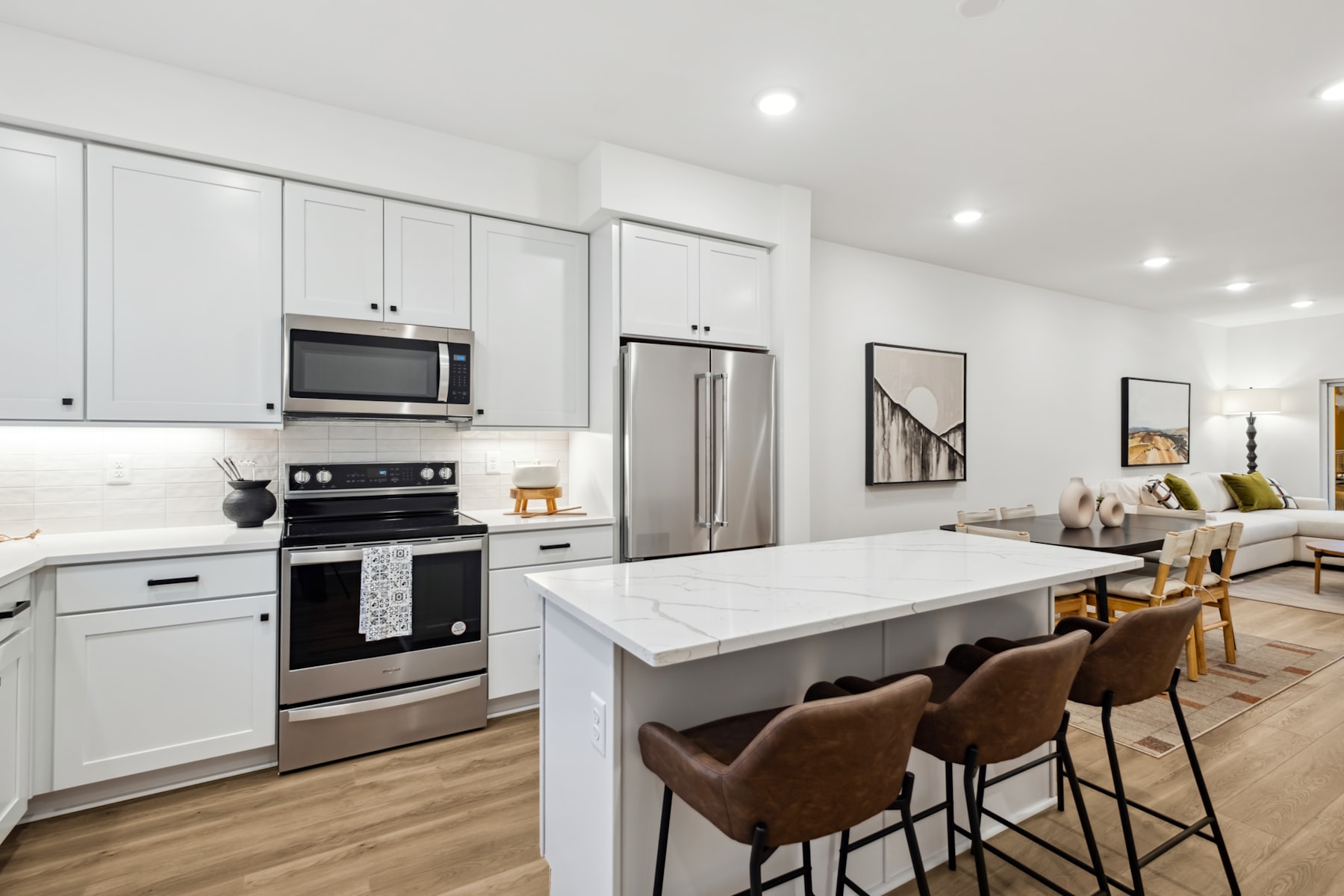 A modern, open-concept kitchen with white cabinets, stainless steel appliances, and a large central island with bar stools, set against a backdrop of a living room area with a couch.