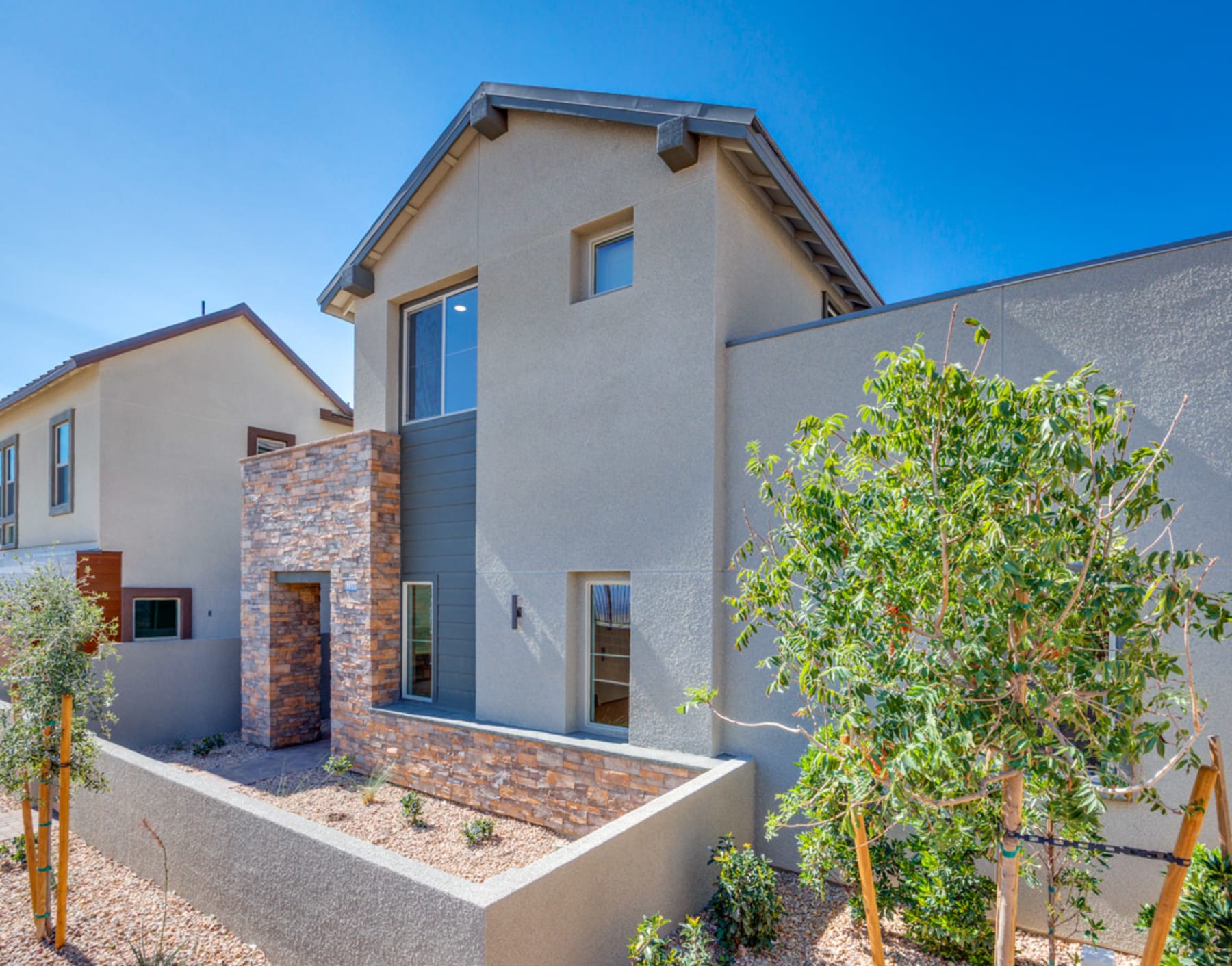 A modern, two-story residential building with a stone facade, surrounded by lush greenery and a paved walkway in the foreground.