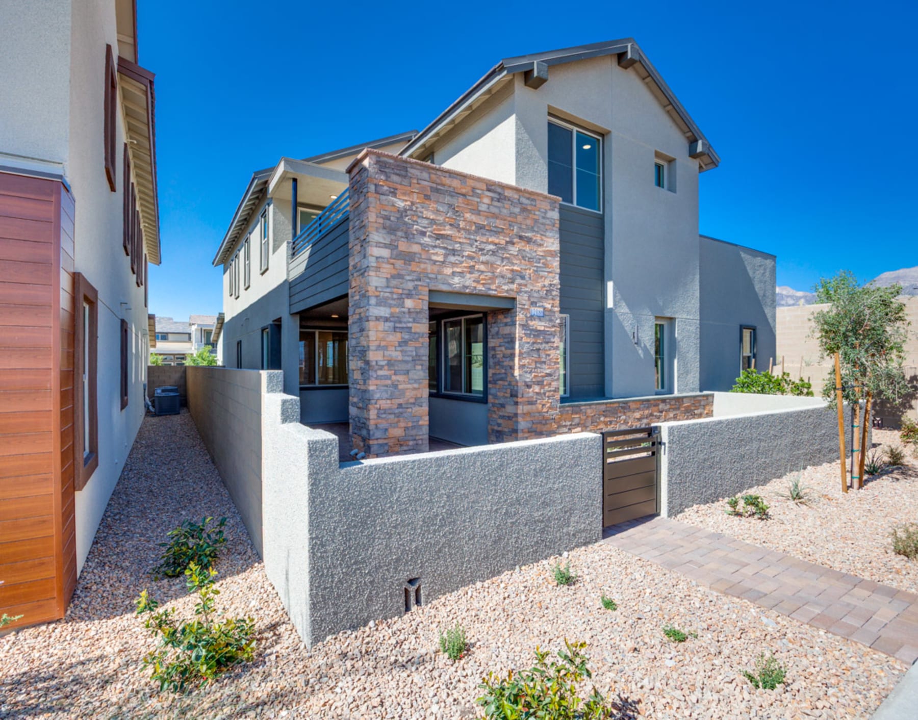 A modern two-story house with a stone exterior, surrounded by a desert landscape and a clear blue sky.