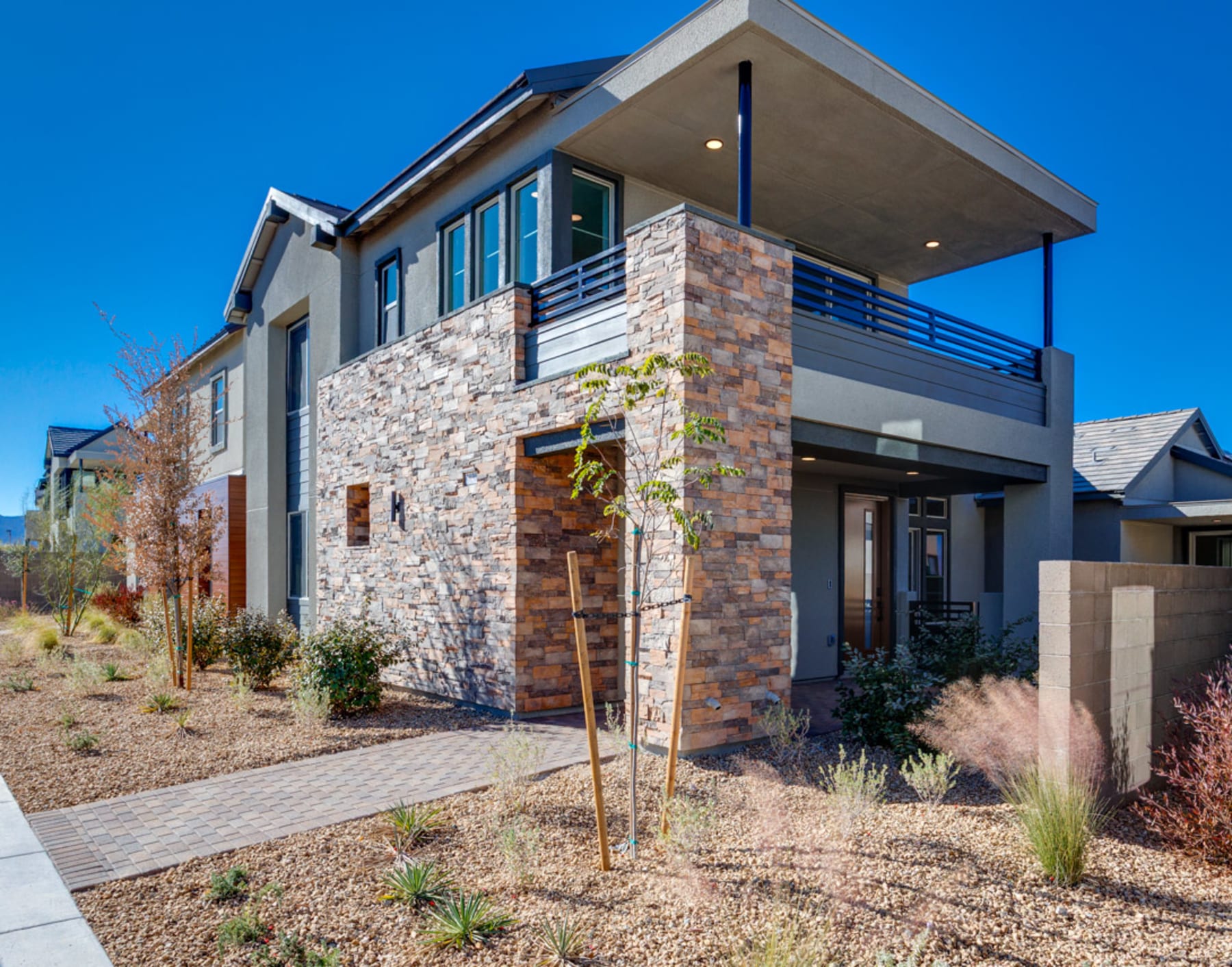 A modern two-story house with a stone exterior, a covered porch, and a landscaped yard with grassy areas and decorative plants in the foreground, set against a clear blue sky.