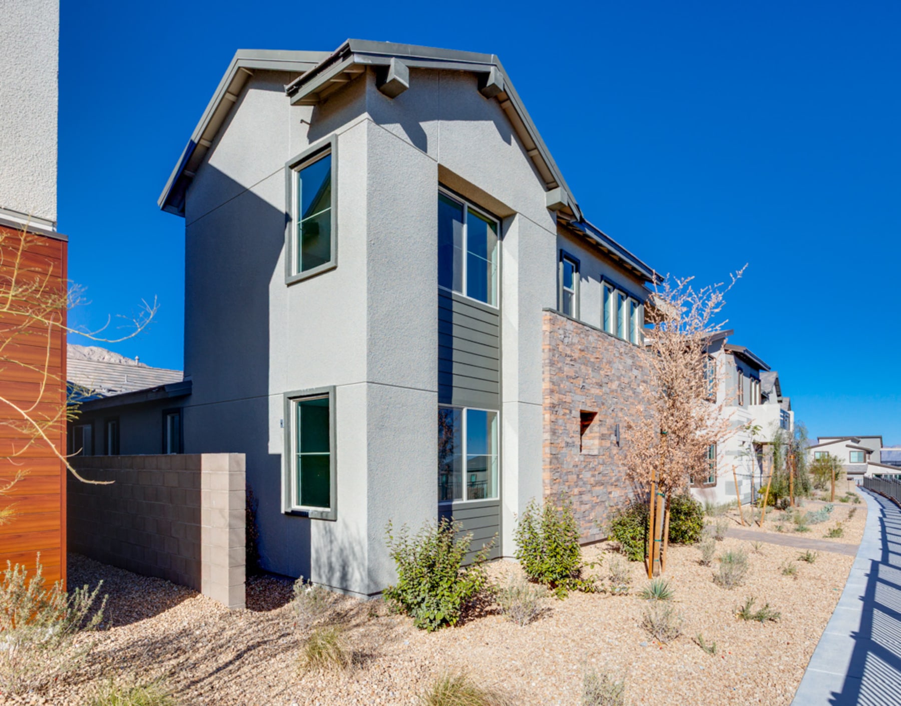 A modern, multi-story residential building with a mix of materials, including stucco and brick, set against a clear blue sky with a landscaped foreground featuring desert plants.