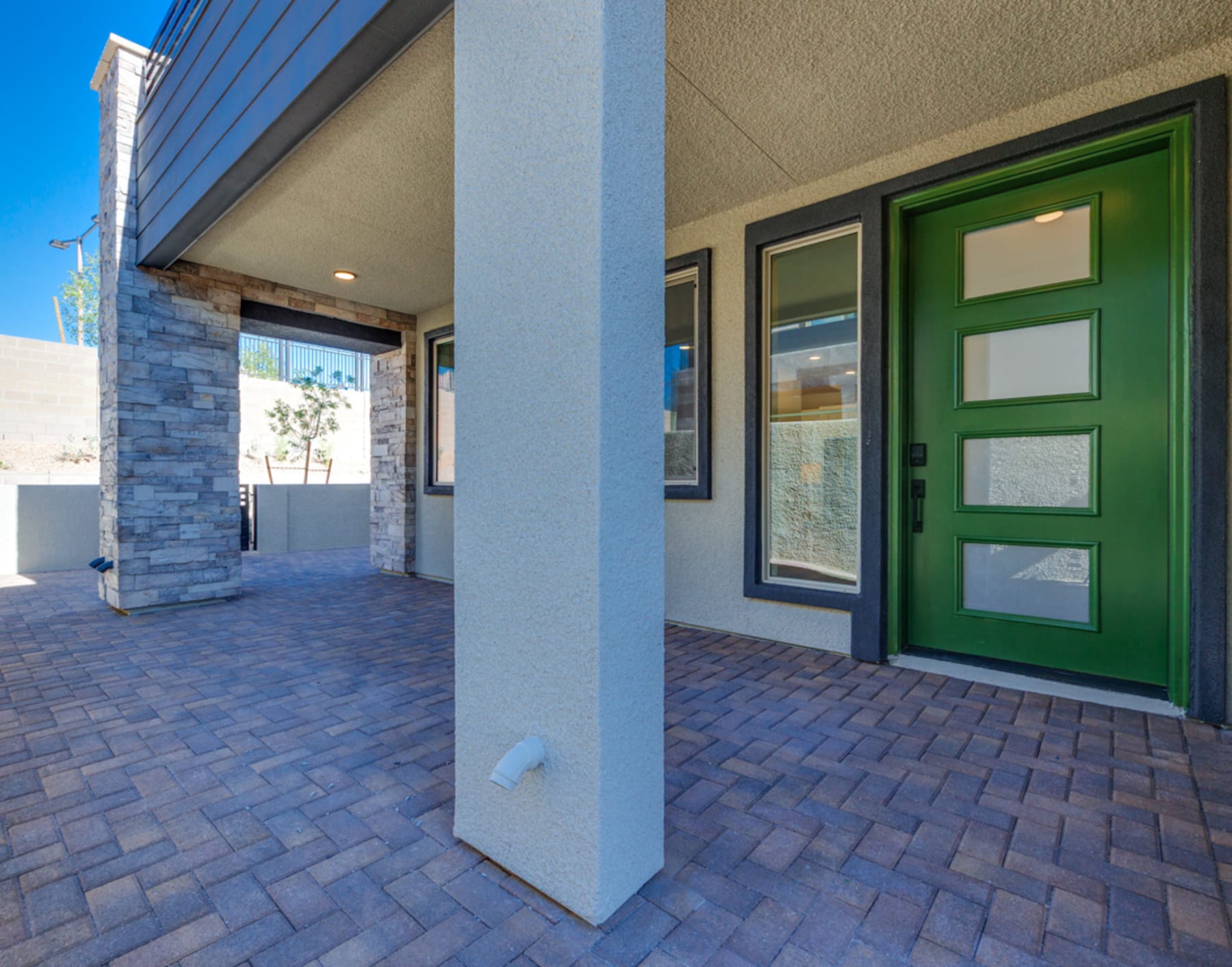 A modern, well-designed entryway with a large green door, brick paving, and a stone exterior, set against a clear blue sky.
