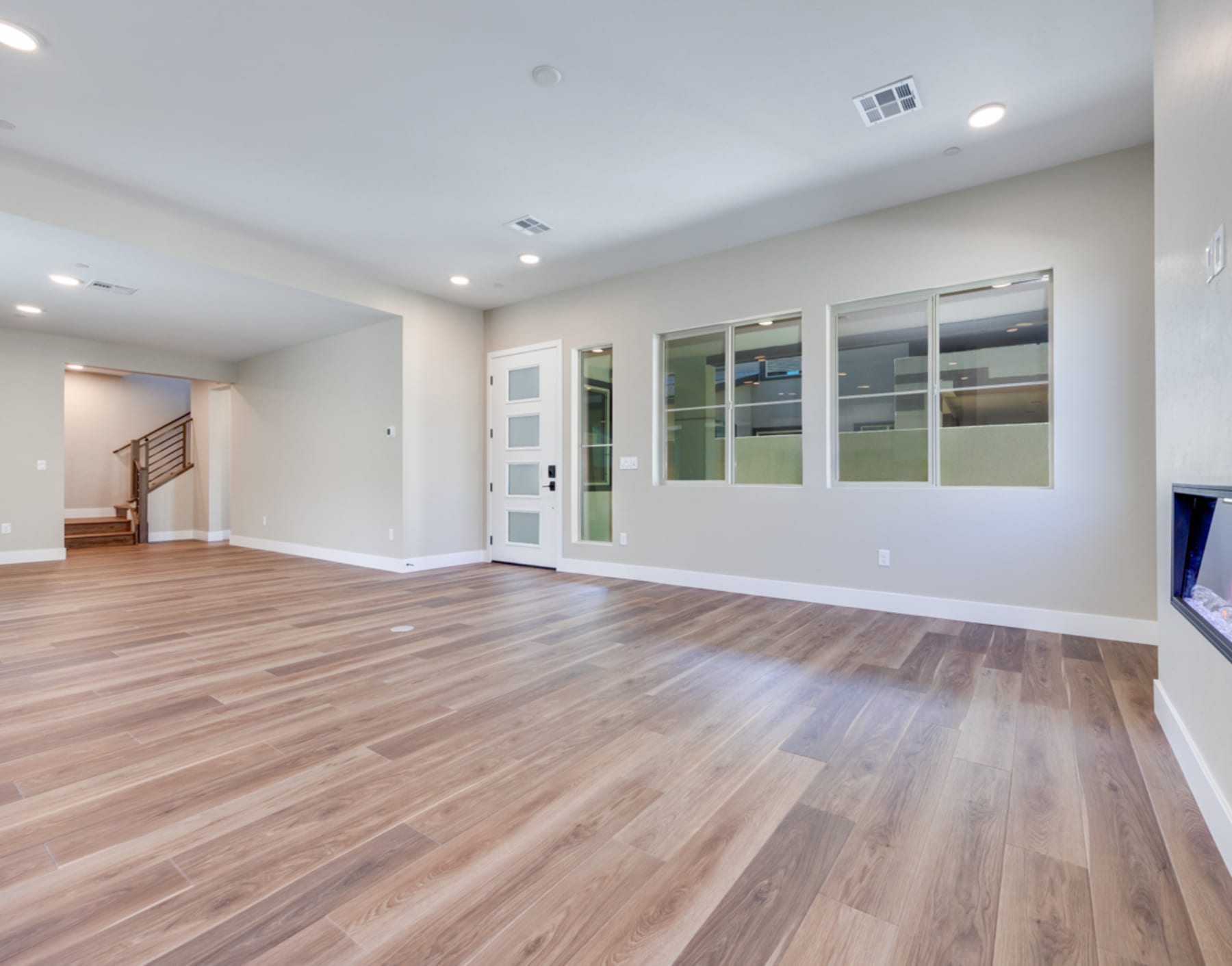 A spacious, well-lit room with hardwood floors, white walls, and large windows, leading to a staircase in the background.