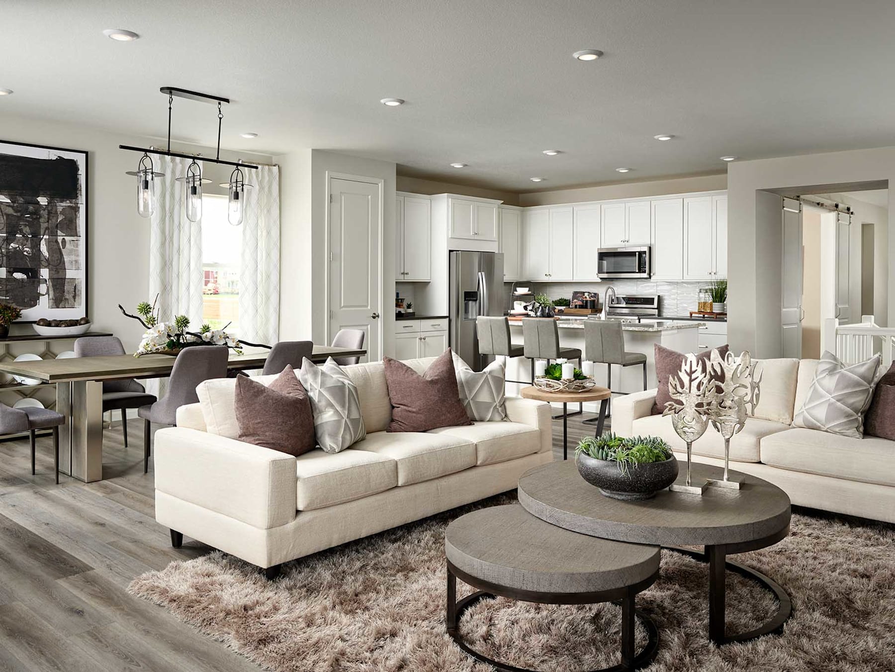 A cozy and modern living room with a plush white sofa, round coffee tables, and an open-concept kitchen visible in the background, all set against a neutral color palette and warm lighting.