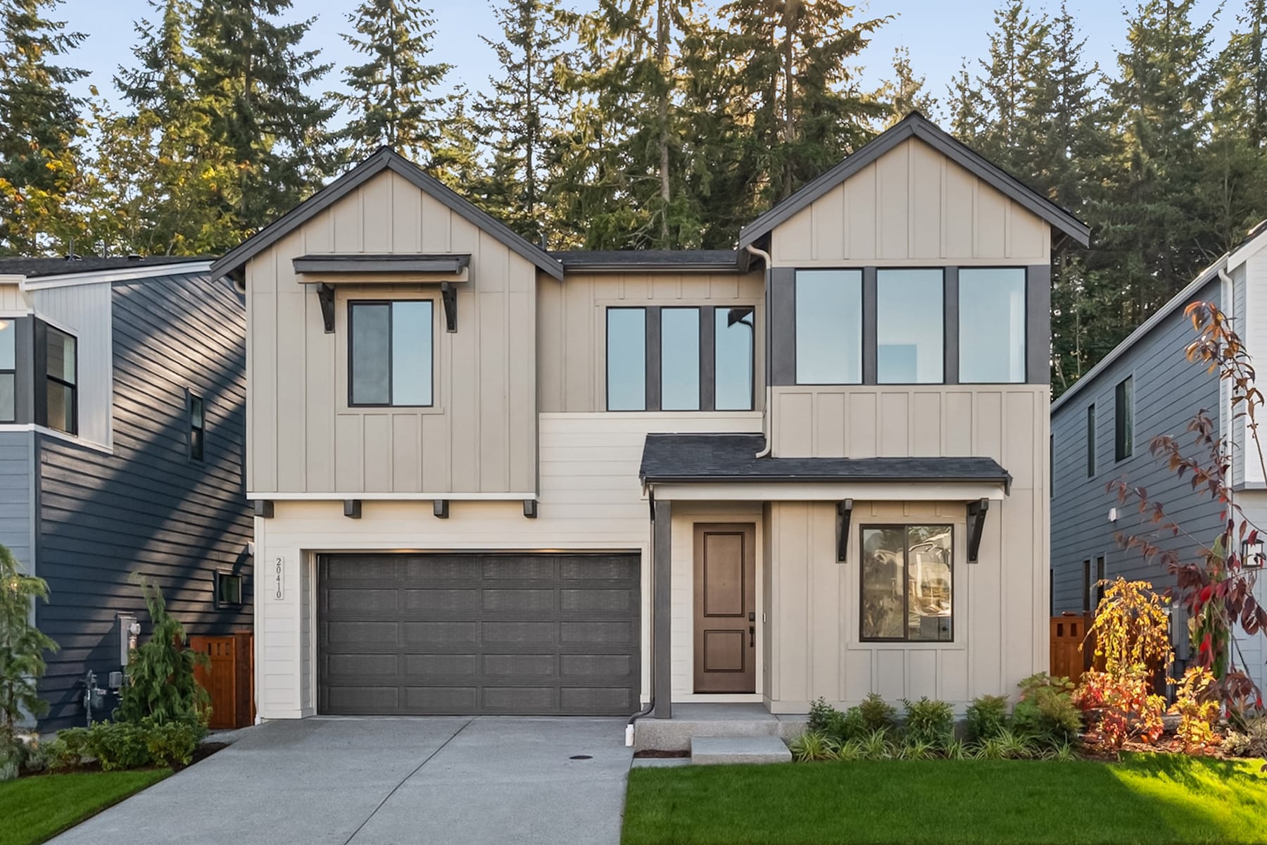 A modern two-story house with a garage, surrounded by lush greenery and tall pine trees in the background.