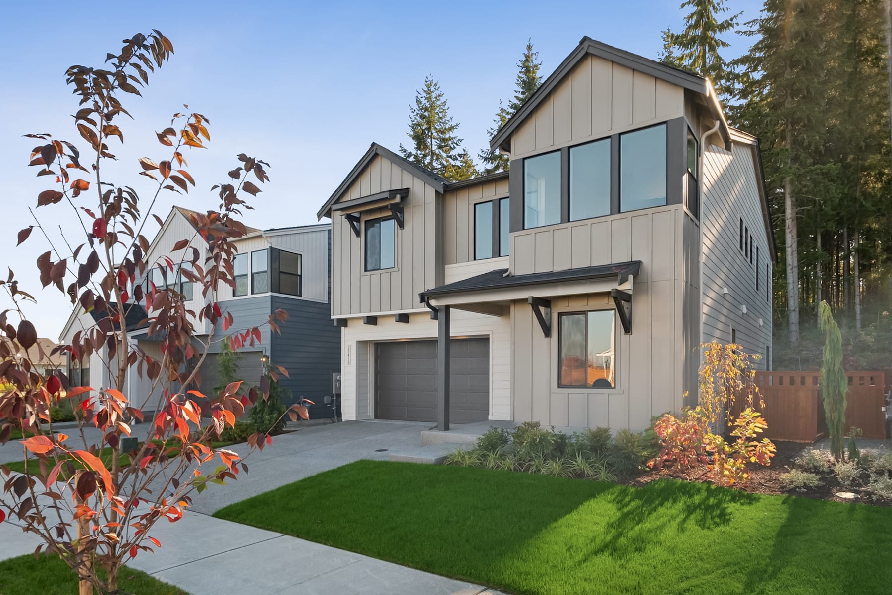 A modern, two-story house with a garage, surrounded by lush greenery and colorful foliage in the foreground, set against a backdrop of tall pine trees.