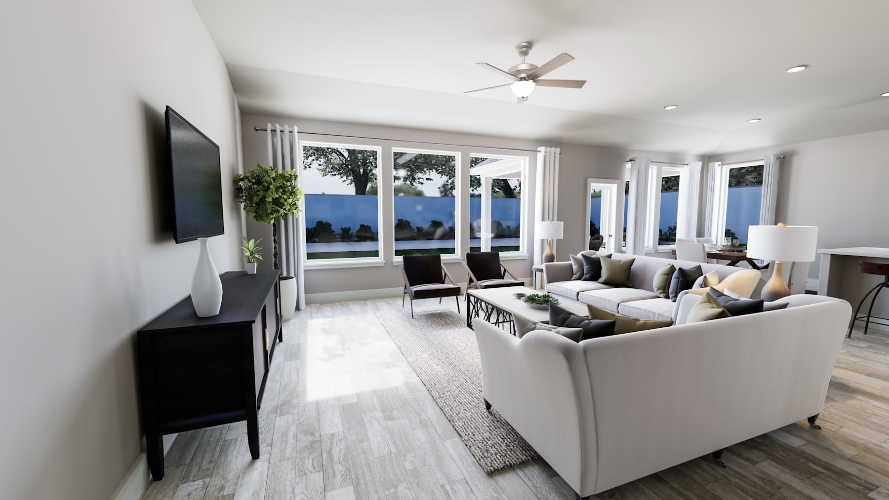 A modern and spacious living room with large windows overlooking a scenic outdoor view, featuring a comfortable seating arrangement, a ceiling fan, and a television mounted on the wall.