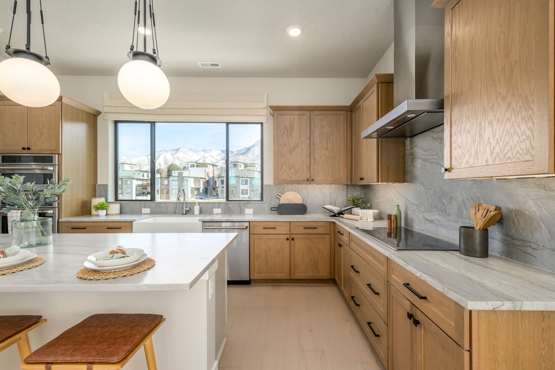A modern, well-lit kitchen with wooden cabinets, white countertops, and pendant lights, overlooking a scenic outdoor view through a large window.