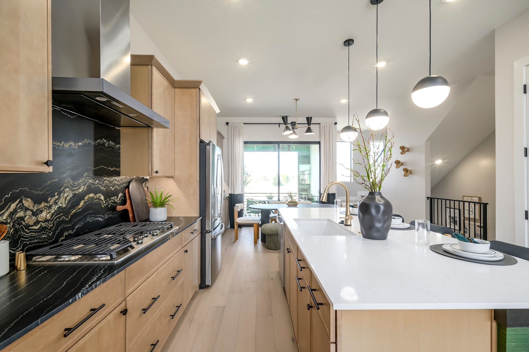 A modern and spacious kitchen with sleek wooden cabinets, a black marble backsplash, and pendant lighting leading into an open living area with a cozy seating arrangement.