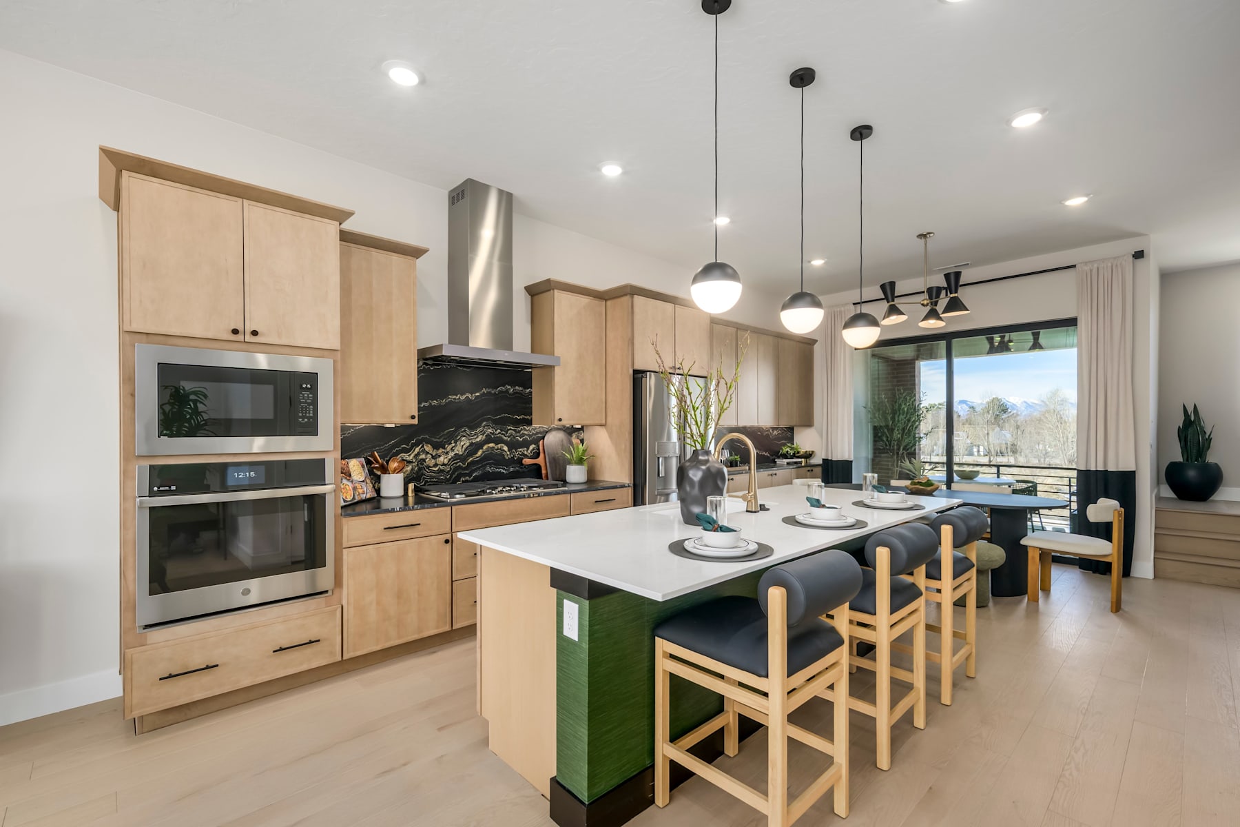 A modern and spacious kitchen with wooden cabinets, a green island, and pendant lights, overlooking a dining area with a large window providing natural light.