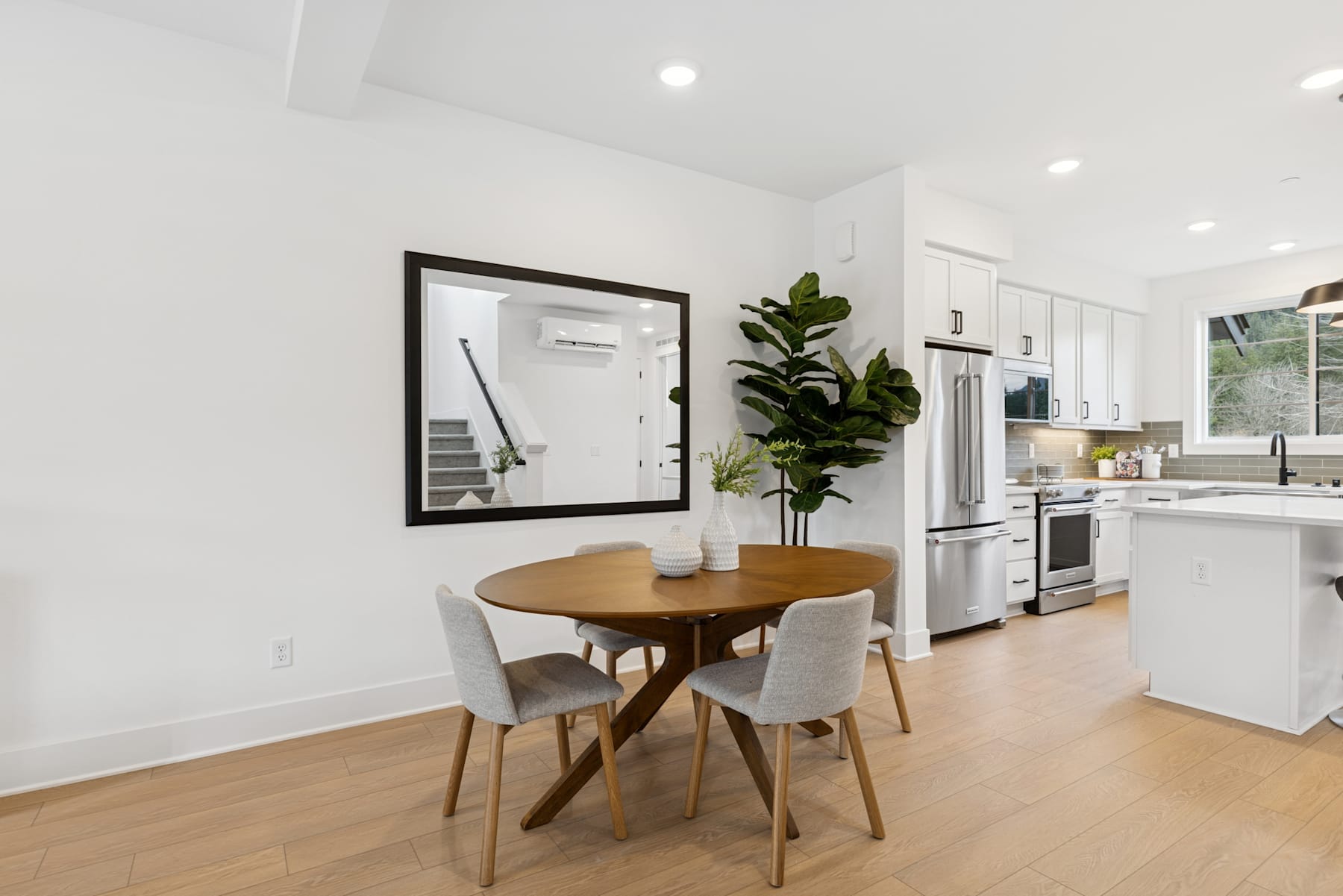 A modern, minimalist kitchen and dining area with a round wooden table, gray chairs, and a large potted plant in the foreground, set against a bright, white-walled background.