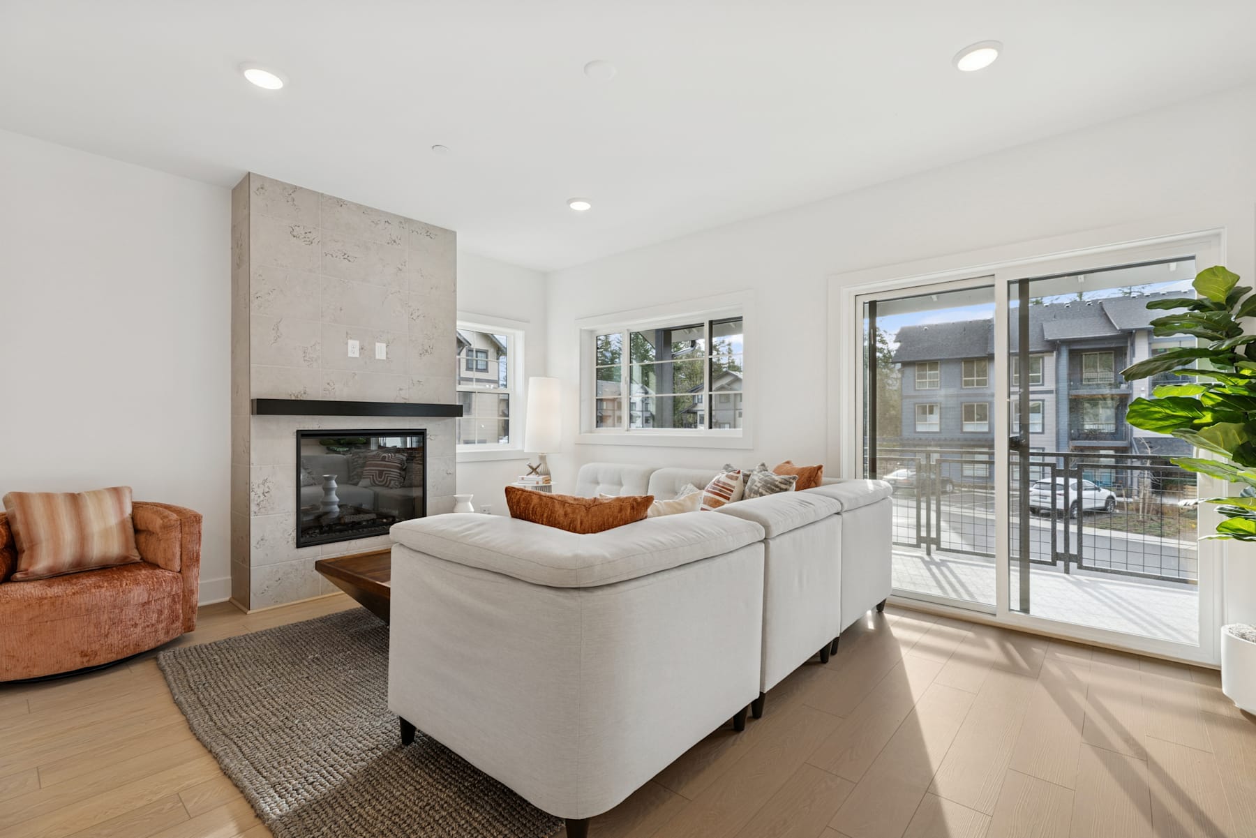 A modern and cozy living room with a white sofa, a fireplace, and large windows overlooking a balcony or patio area.