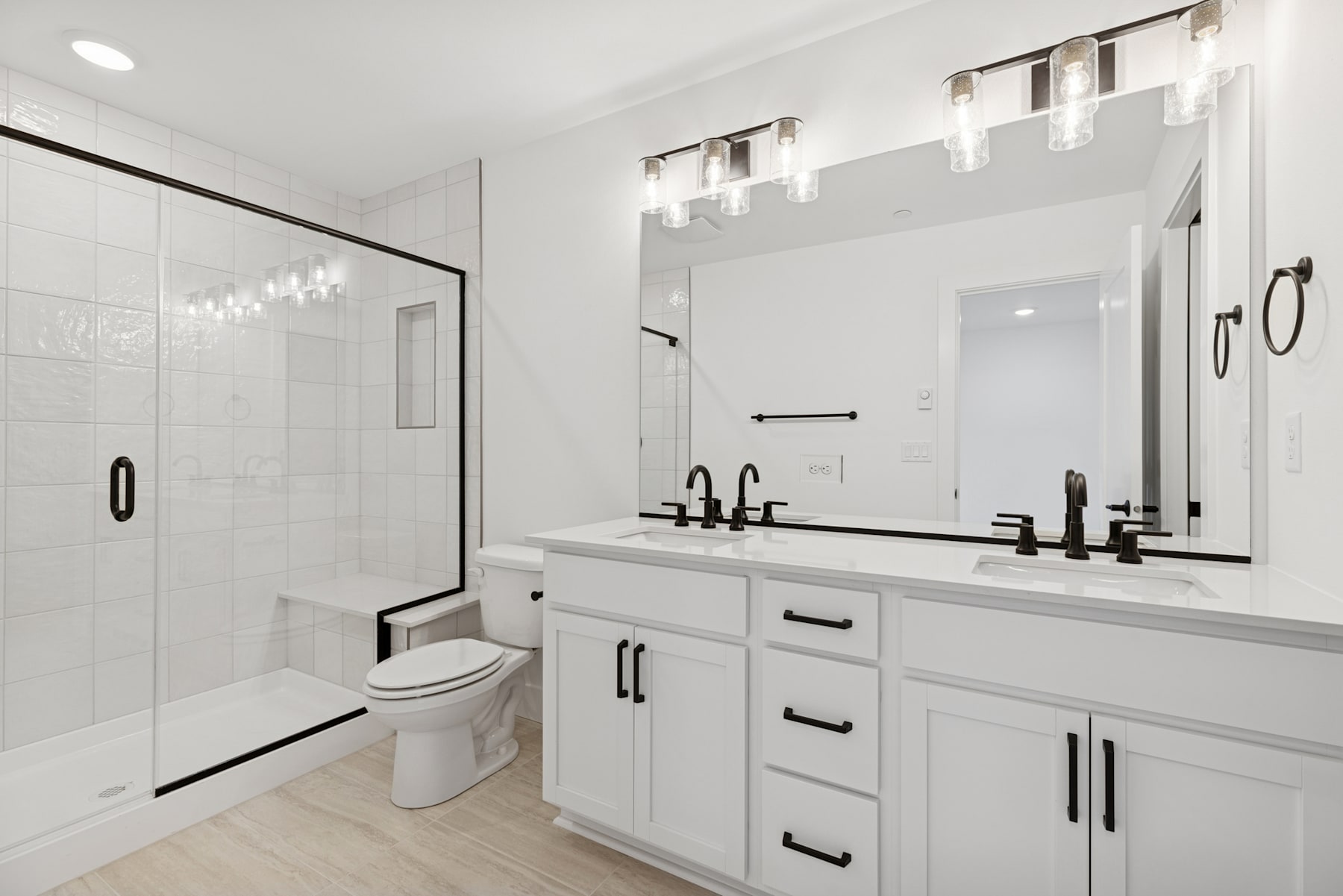 A modern and minimalist bathroom with white cabinets, a double vanity, and a large mirror with overhead lighting. The room features a tiled shower enclosure and a toilet.