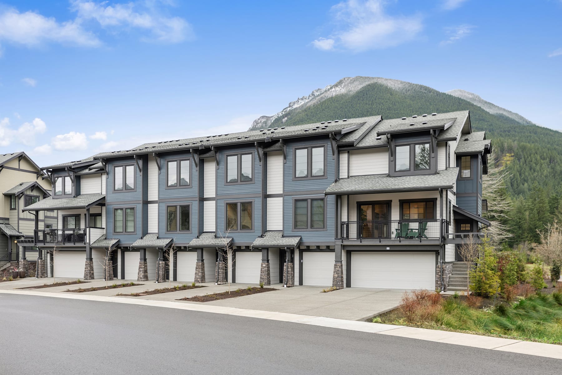 A multi-story residential building with attached garages sits in the foreground, surrounded by lush greenery and a mountainous landscape in the background.