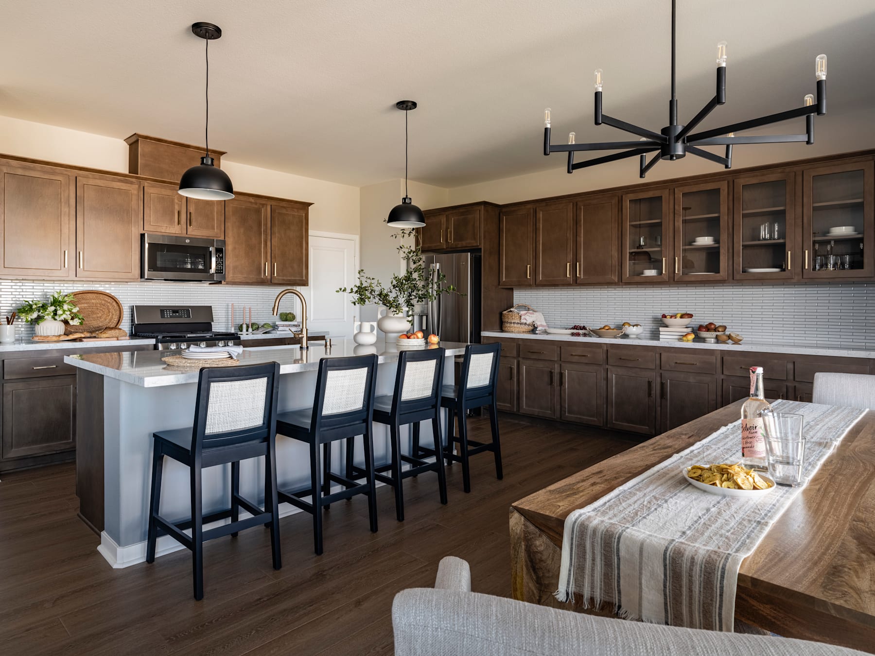 A modern and spacious kitchen with wooden cabinets, a large island with bar stools, and a chandelier overhead, set against a backdrop of a dining area with a table and chairs.