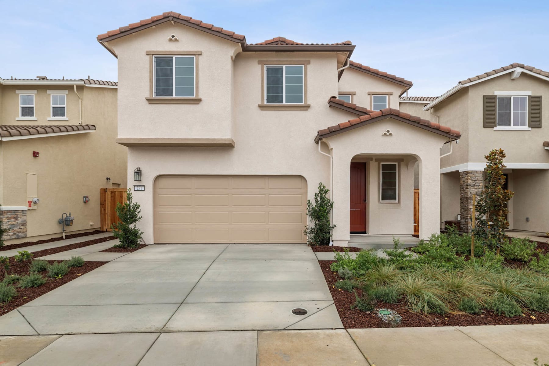 A two-story Mediterranean-style house with a red tile roof, a paved driveway, and a well-landscaped front yard with various plants and shrubs.