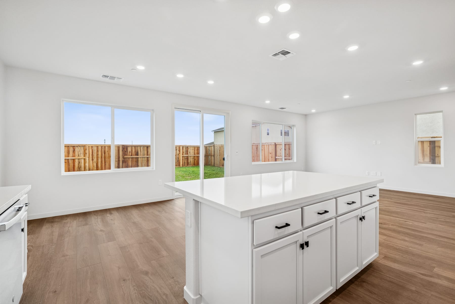 A bright and spacious kitchen with white cabinets, a large countertop, and wooden flooring, surrounded by large windows overlooking a backyard.