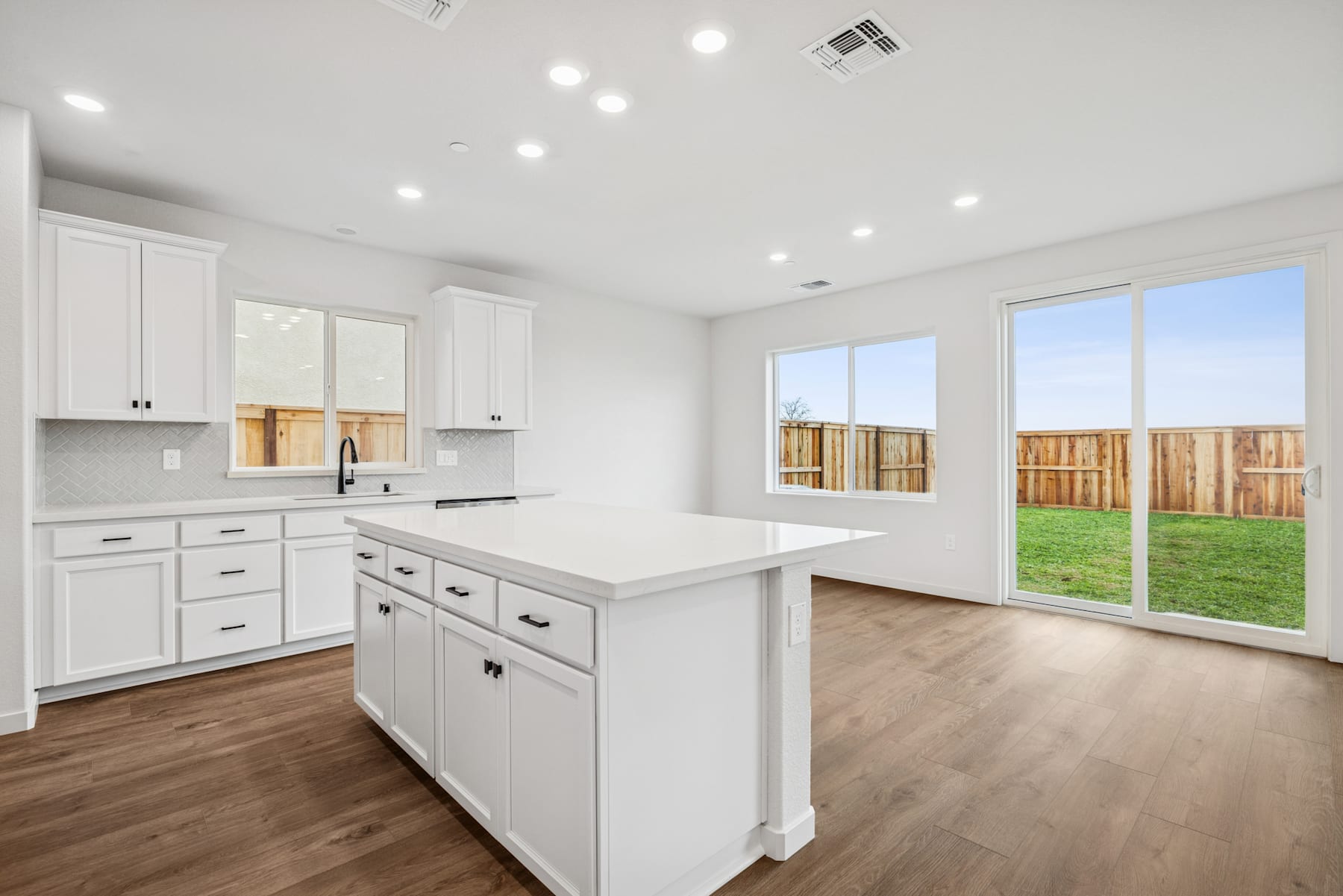 A modern, bright kitchen with white cabinets, a central island, and large windows overlooking a grassy backyard.